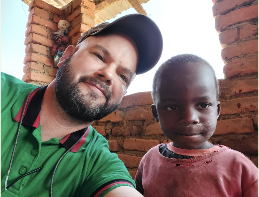 A man wearing a green polo shirt, gray cap, and with a beard takes a selfie with a young boy with short hair and a red shirt in front of a brick wall with open sky visible in the background.