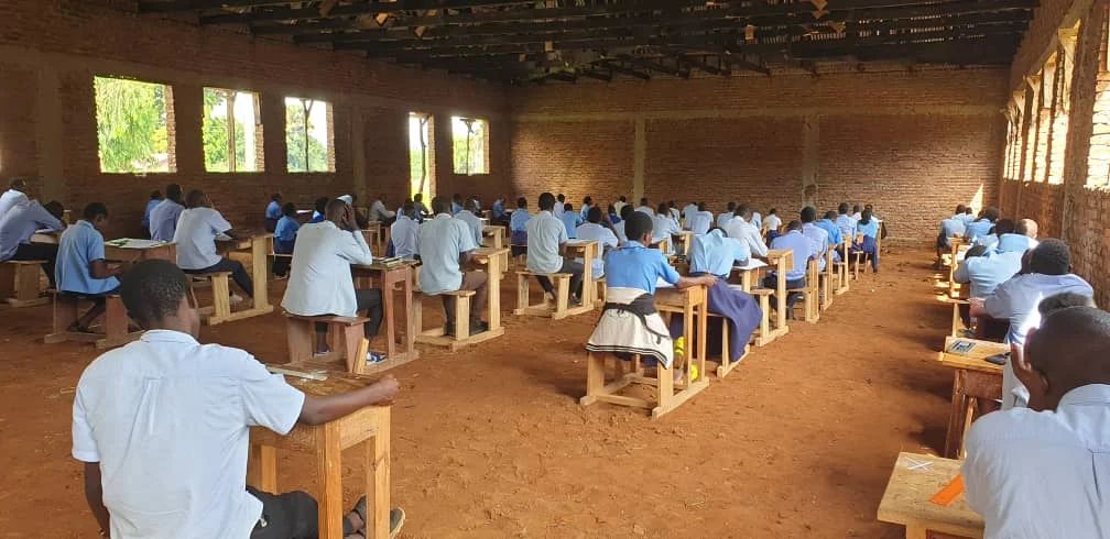 Students in white and blue uniforms seated at wooden desks taking an exam in a classroom with brick walls and windows.