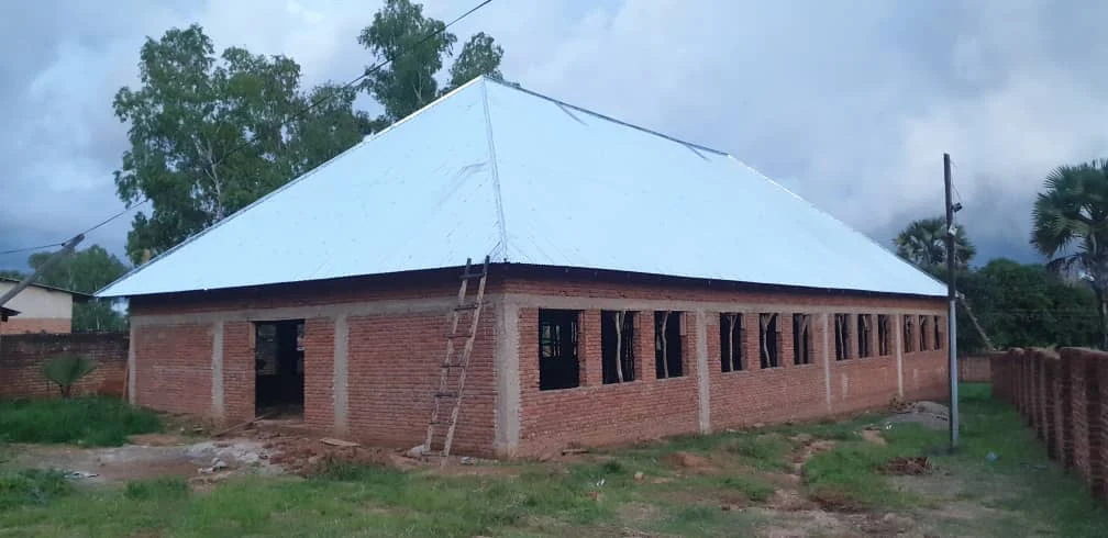 Brick building under construction with a metal roof, ladder leaning against it, and a grassy area in the foreground.