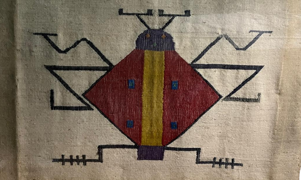 A textile or embroidered depiction of a geometric bug or insect with a diamond-shaped red and yellow body, blue squares, and black antennae and legs on a beige fabric background. New Palm Africa Huckleberry Fox Foundation