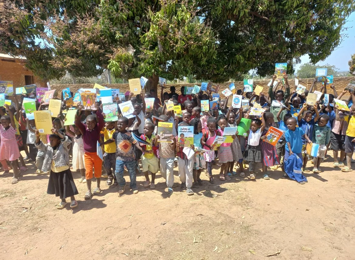 Group of children outdoors holding up children's books under a large tree.
