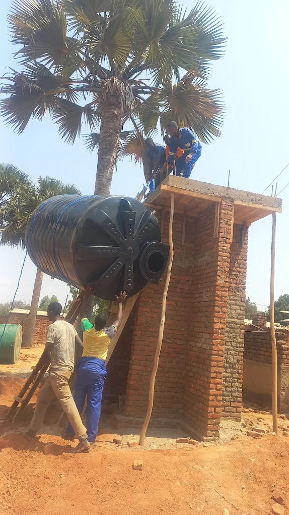 Two men on a brick water tower, one on top and one below, installing a large black water tank with two other men outside helping, in a rural setting with a tall palm tree in the background.