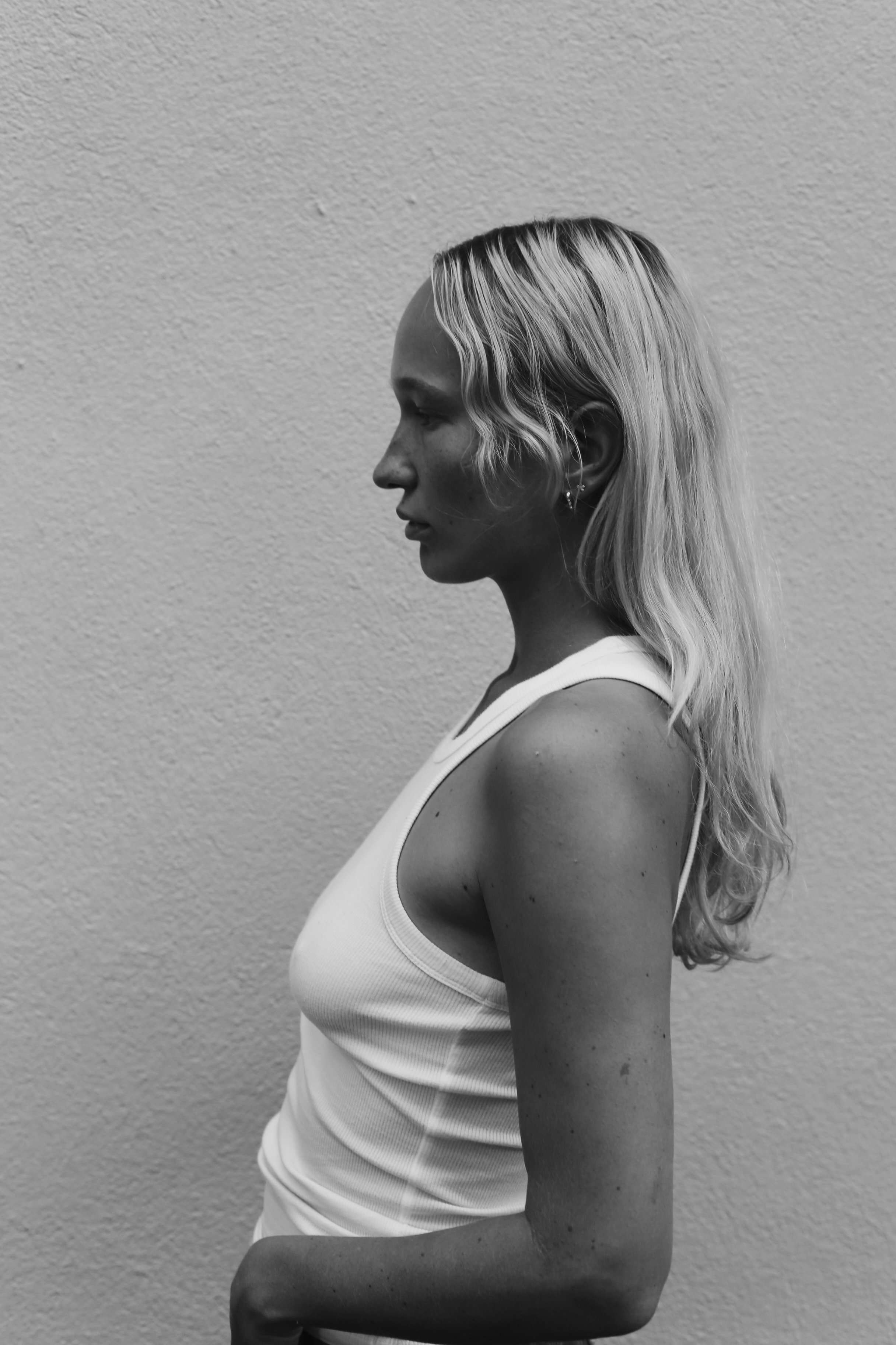 Black and white photo of a woman with light wavy hair in profile, wearing a sleeveless white top, standing against a textured wall.