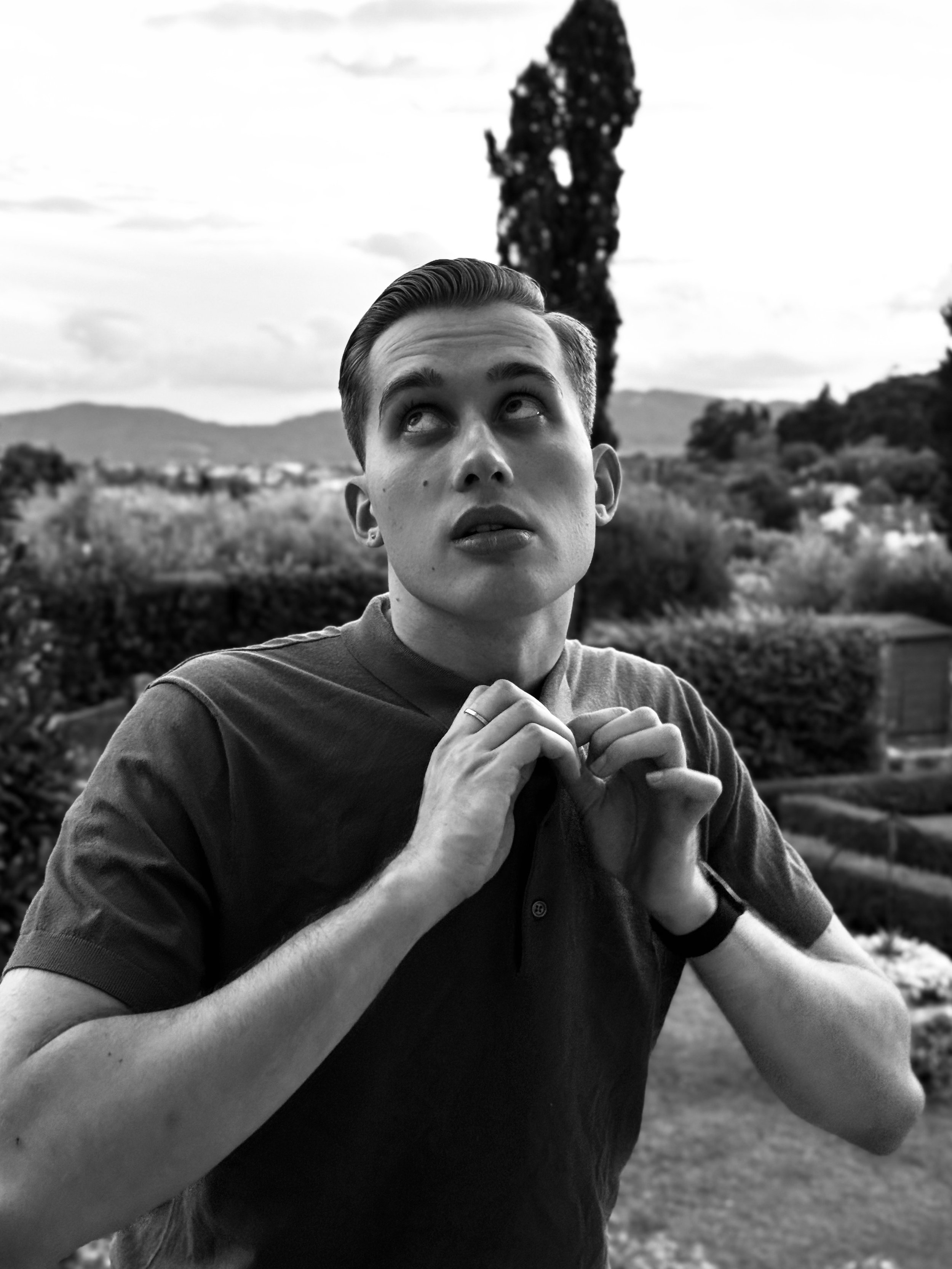 A young man with styled hair and a serious expression, looking up and holding the collar of his shirt, outdoors with trees and mountains in the background.