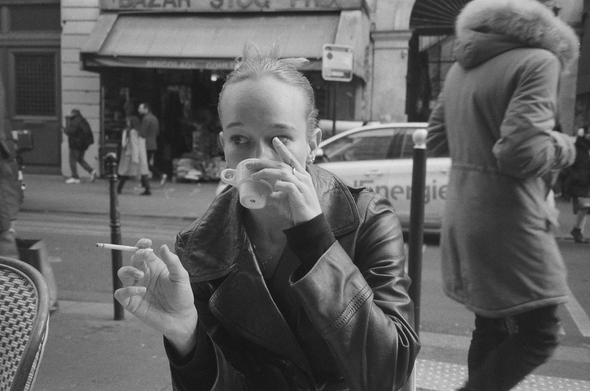 A woman sitting at an outdoor café, drinking from a coffee cup and holding a cigarette, with people walking by on the street behind her.