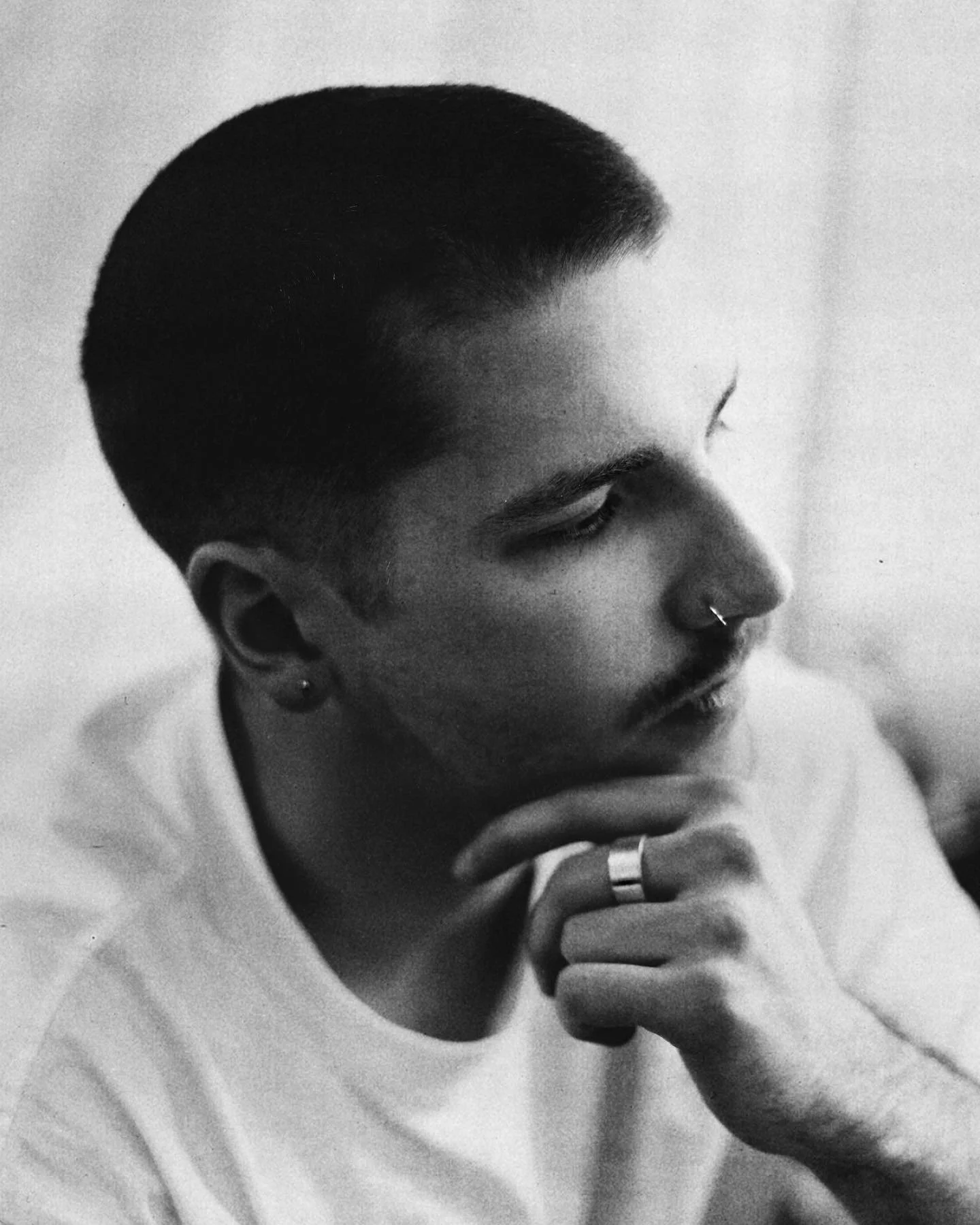 Black and white portrait of a young man with short hair, nose piercings, and rings on his fingers, resting his chin on his hand and looking downward.