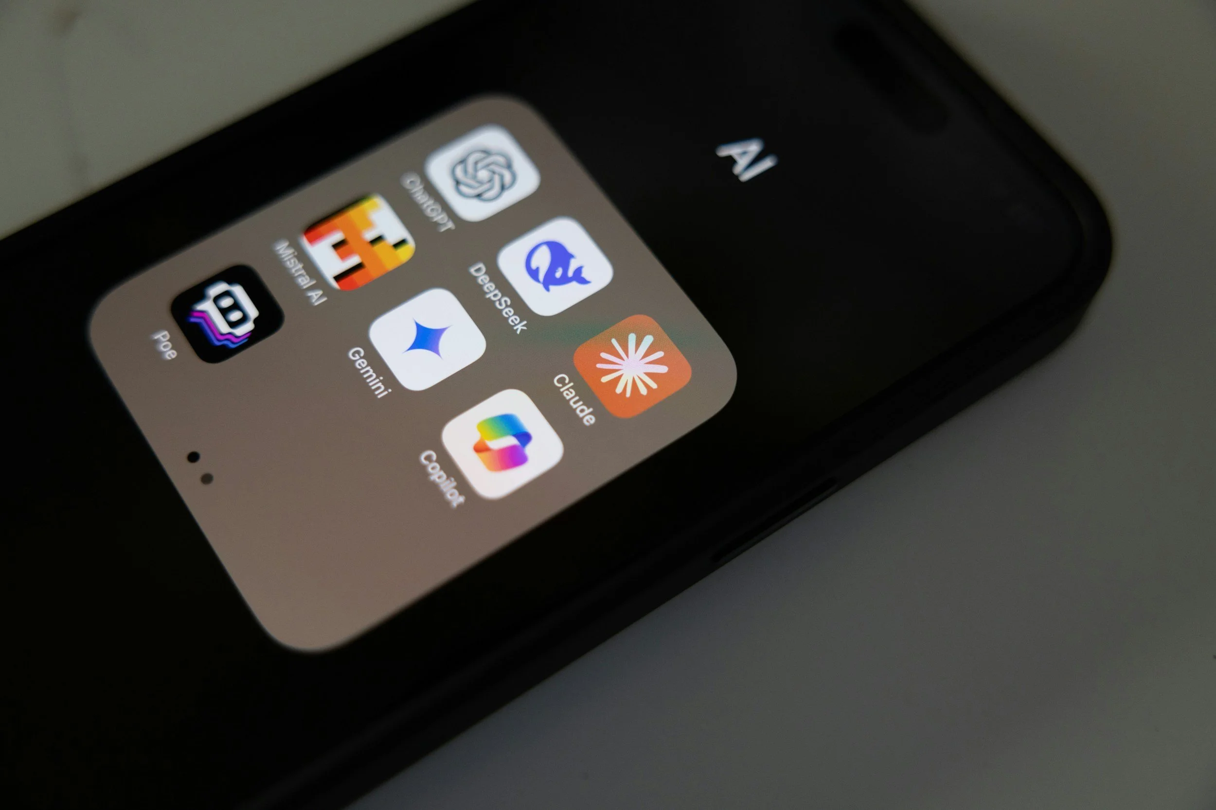 Close-up of an iPhone screen showing a folder with six app icons, including DeepSeek, Poe, Gemini, Claude, and Google, on a dark background.