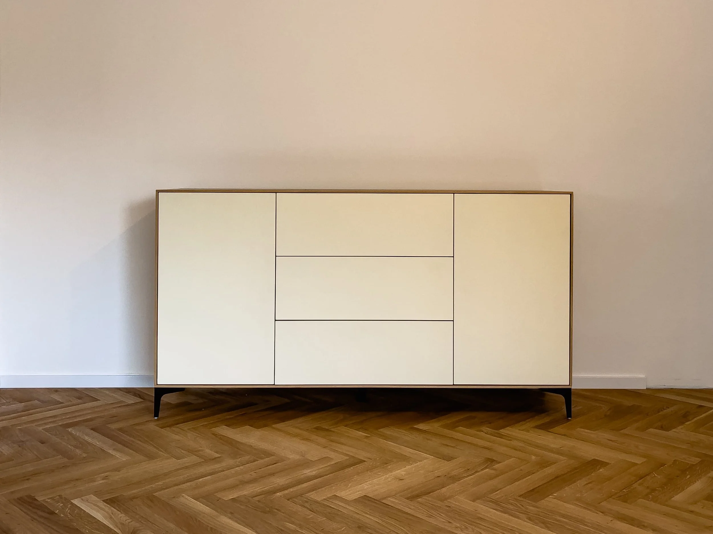 Ein weißer moderner Sideboard mit schwarzen Beinen steht vor einer weißen Wand auf Holzboden.