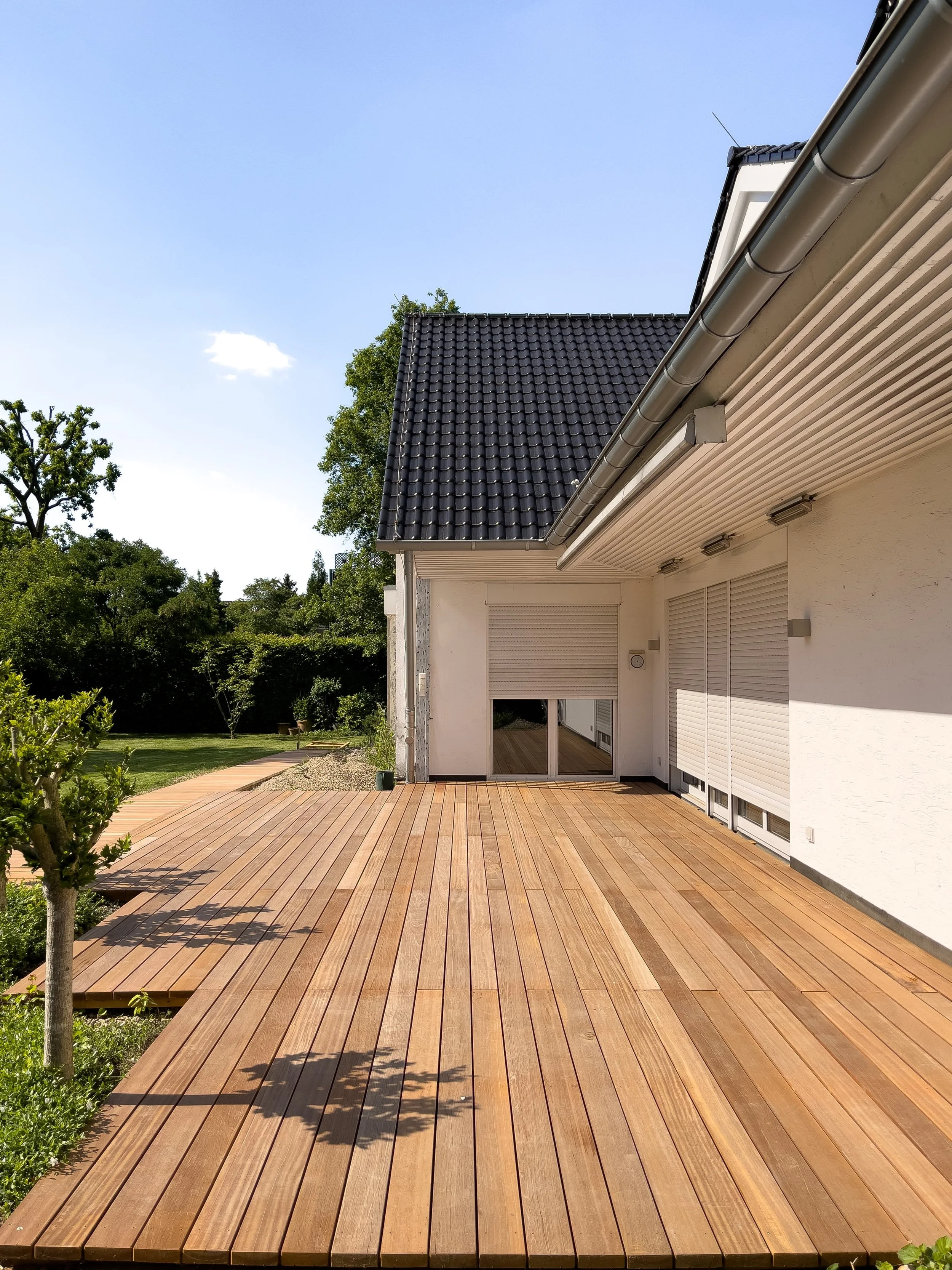 Ein moderner Balkon mit Holzdielen, teilweise beschattet von einem Dachüberstand, und einem weißen Haus mit Rollläden. Im Hintergrund sind Bäume und ein blauer Himmel sichtbar.