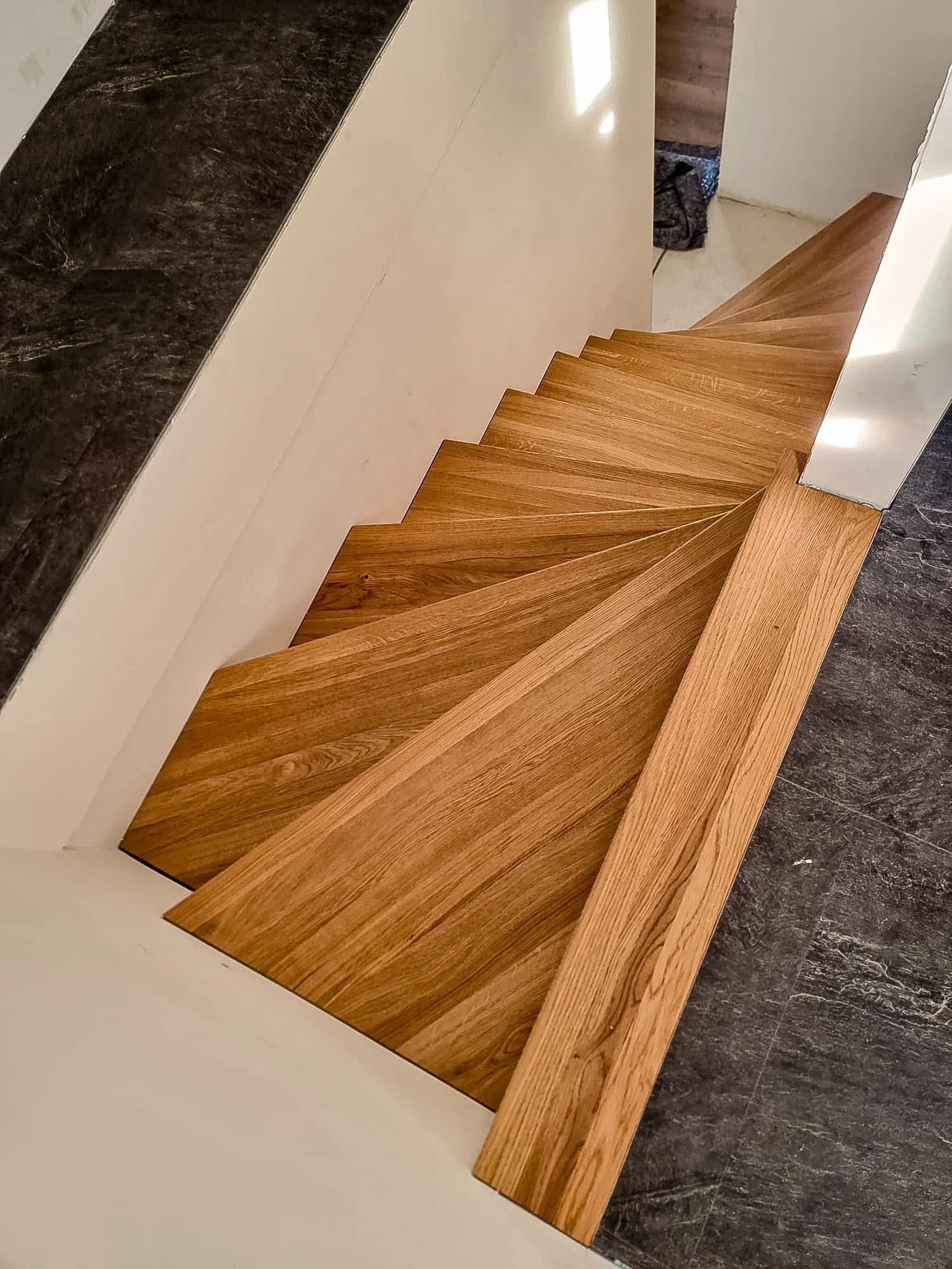 Holztreppe mit schrägem Design, die in eine Raumecke führt, umgeben von weißen Wänden und dunklem Bodenbelag.