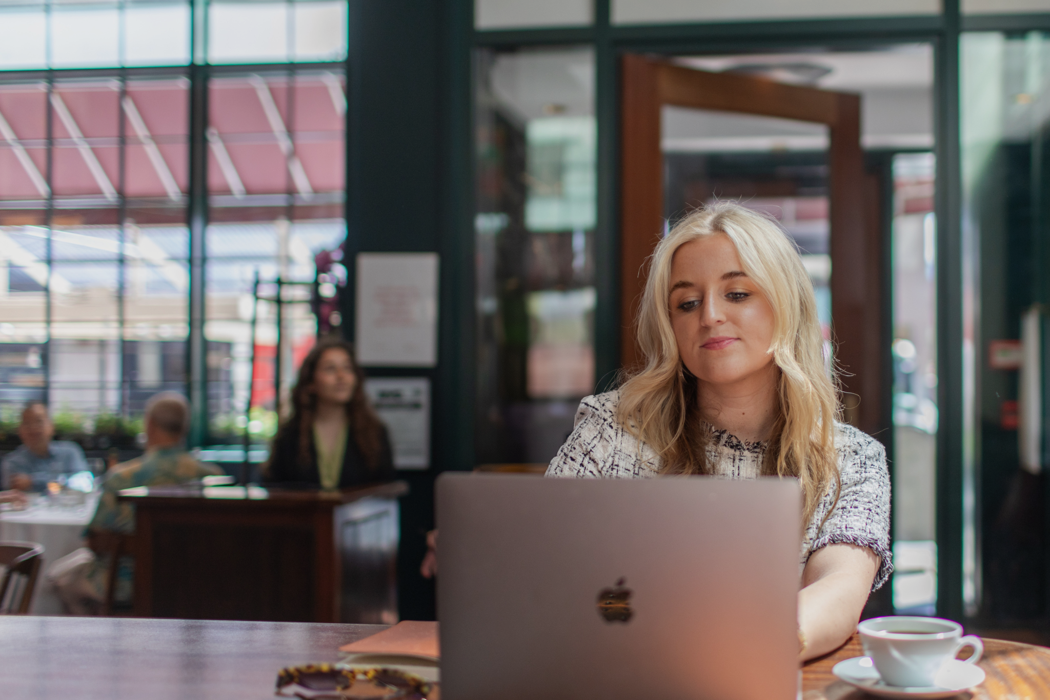A blonde woman working on a laptop at a table in a busy cafe with other people in the background.