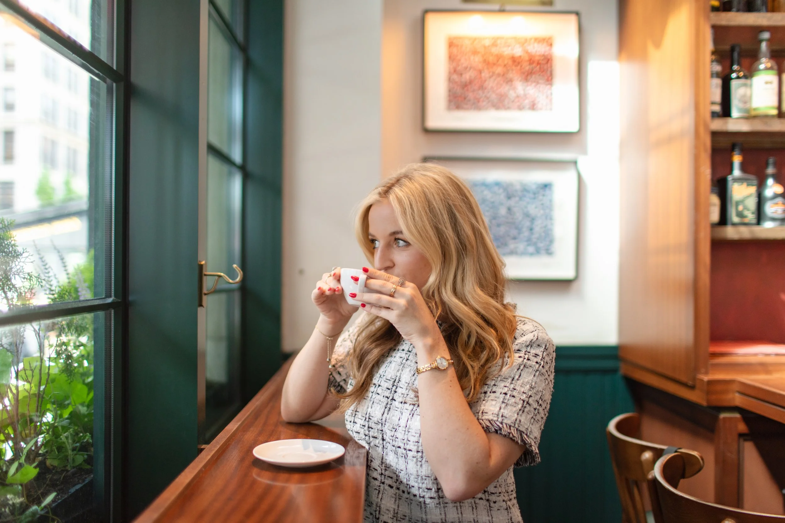 A woman with red hair and light skin, wearing a patterned top and jewelry, sitting at a wooden counter in a cafe, drinking from a white mug, with greenery visible through a window to her left and artwork on the wall behind her.