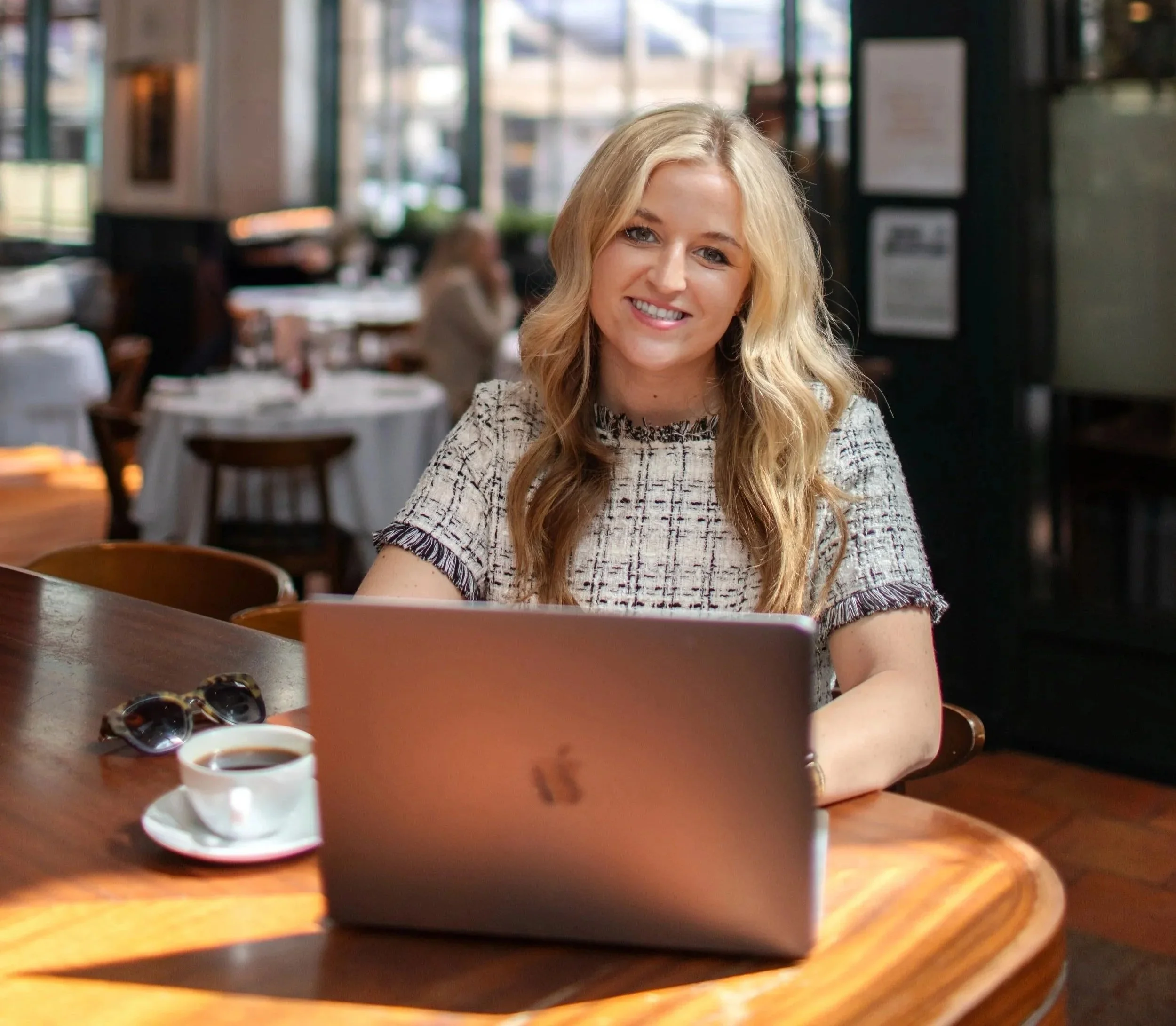 A woman with blonde wavy hair smiling while sitting at a wooden table in a restaurant. She is wearing a light-colored patterned top. On the table, there is a laptop, a cup of coffee, and sunglasses. In the background, there are large windows with daylight streaming in, and other diners visible.