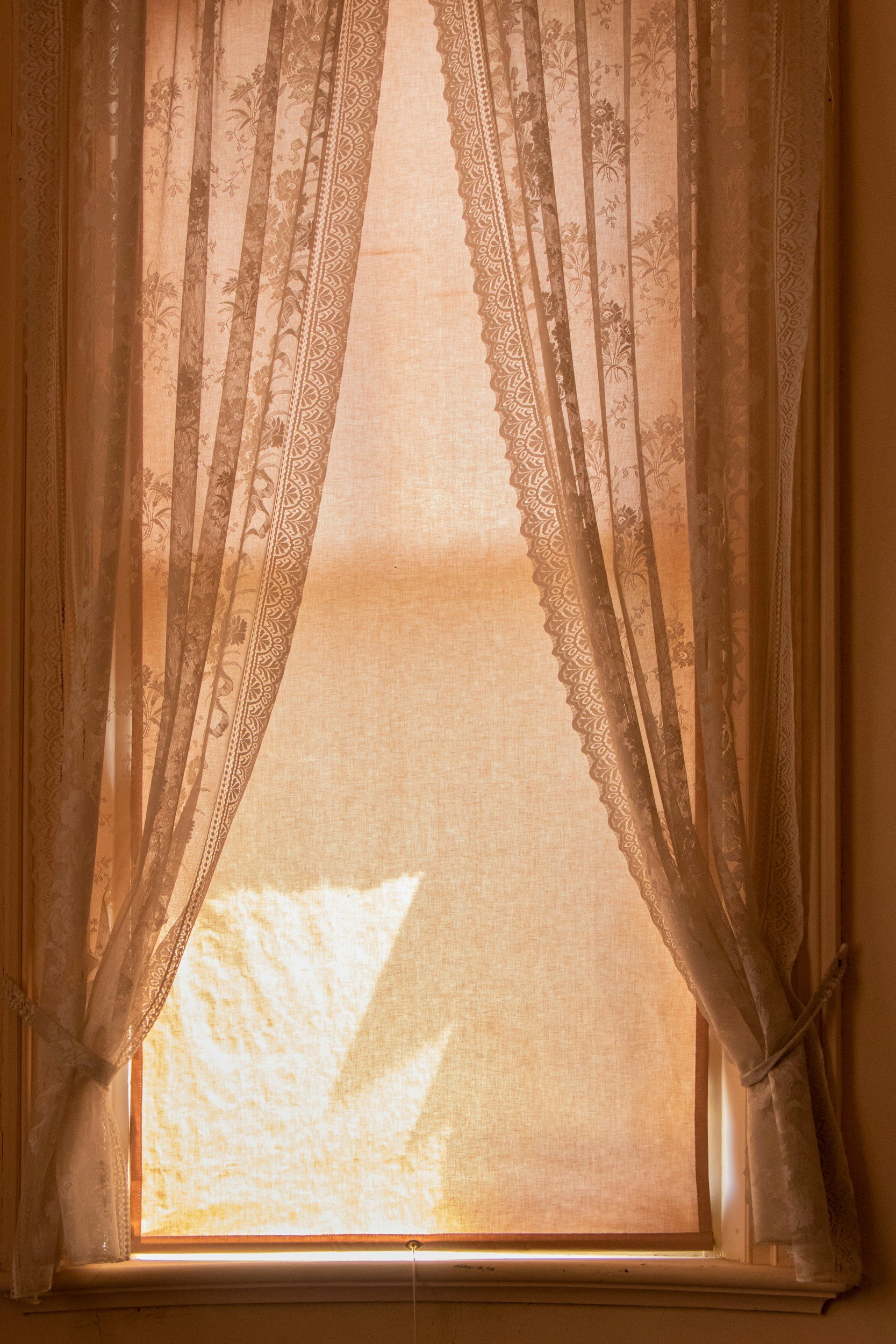 A window with beige lace curtains partially drawn, allowing sunlight to shine through.