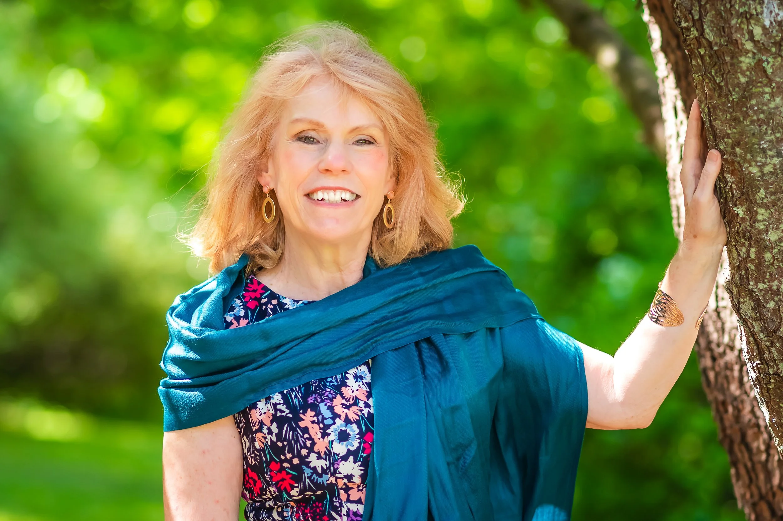 A woman with shoulder-length blonde hair smiling outdoors, wearing floral dress, gold hoop earrings, a blue scarf draped over her shoulders, and a bracelet, standing next to a tree with green foliage in the background.