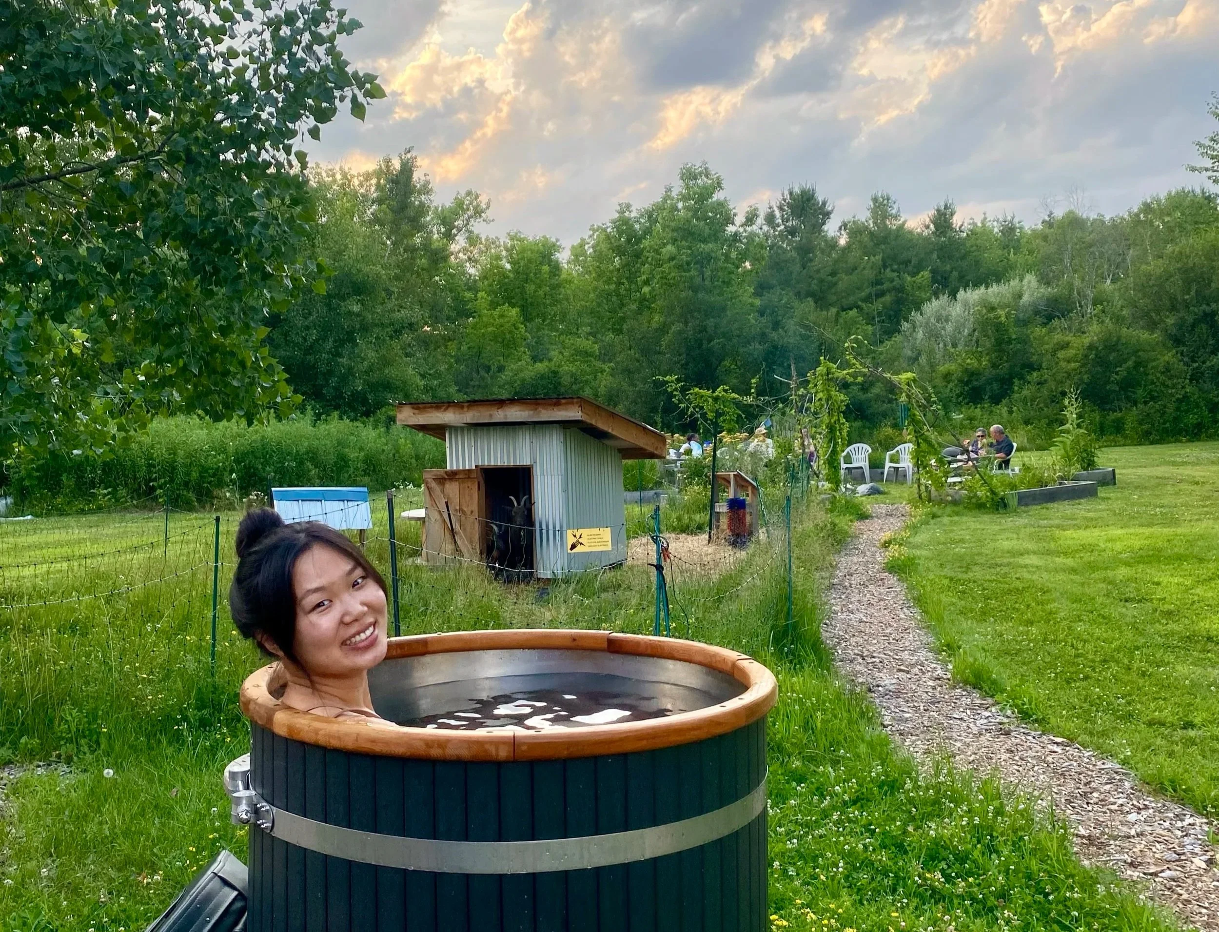 A young woman smiling while sitting in a wooden hot tub in a green outdoor garden at sunset. In the background, there are trees, a small chicken coop, a blue container, and people sitting on white plastic chairs around a table.