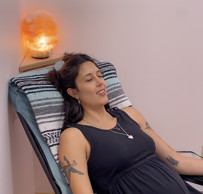 A woman with dark hair and tattoos relaxing on a reclining chair with a striped pillow, a black sleeveless dress, and closet shelves visible behind her with a salt lamp on a small wooden shelf.