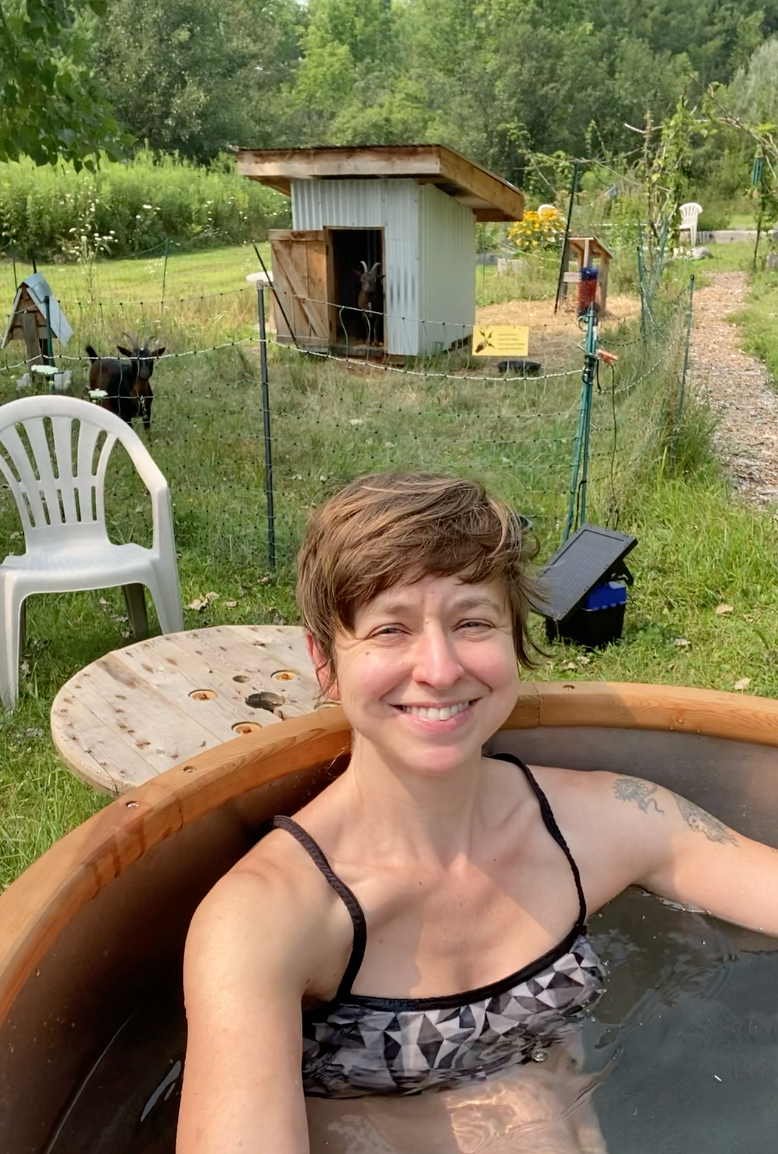 A woman smiling while relaxing in a wooden hot tub outdoors near a fenced area with goats and a shed, in a lush green setting.