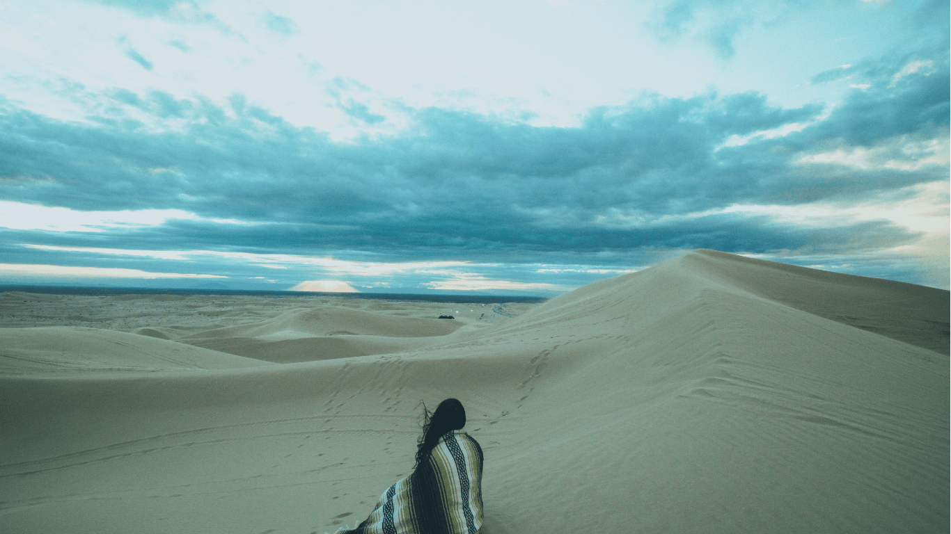 Woman sitting on sand dunes in a reflective moment, representing personal growth, identity shift, and overcoming old insecurities.