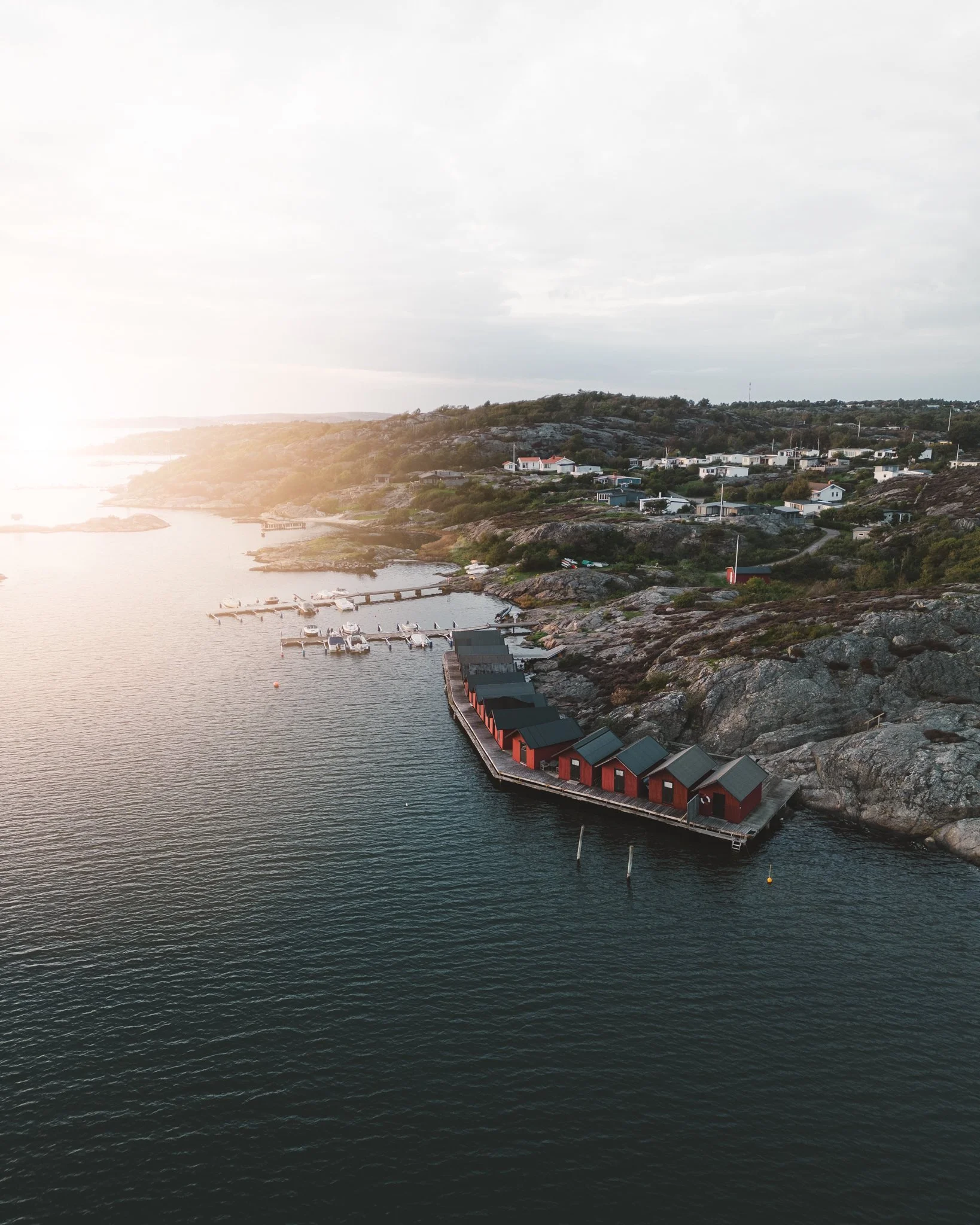 Vy över en småbåtshamn med röda bodar längs vattnet, omgiven av klippor och ett bostadsområde på en kullig landsväg, under ett molnigt himmel.