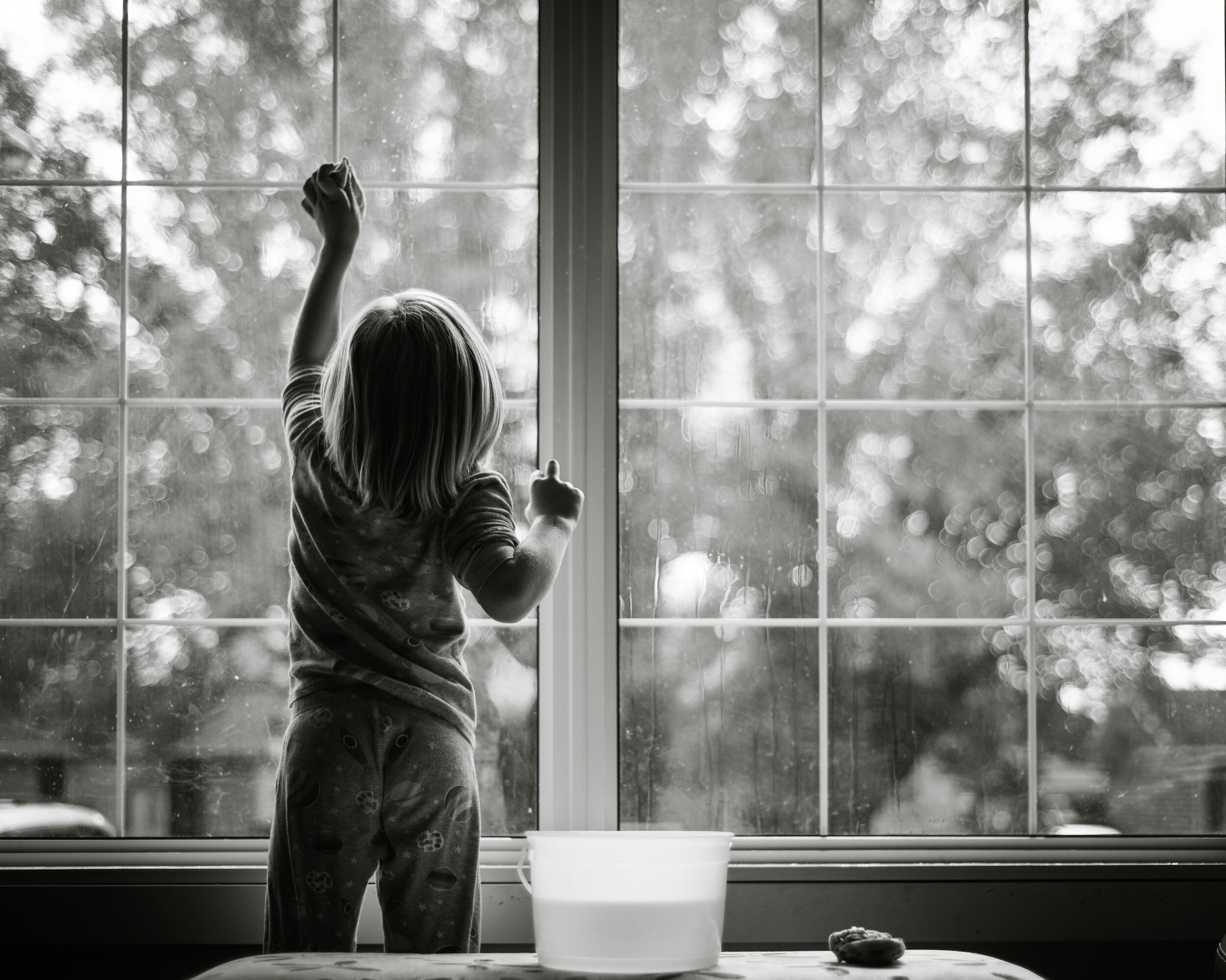 A young girl with shoulder-length hair in pajamas, standing by a large window, looking outside and reaching up with her right hand. There is a white bowl and a small object on the table in the foreground.
