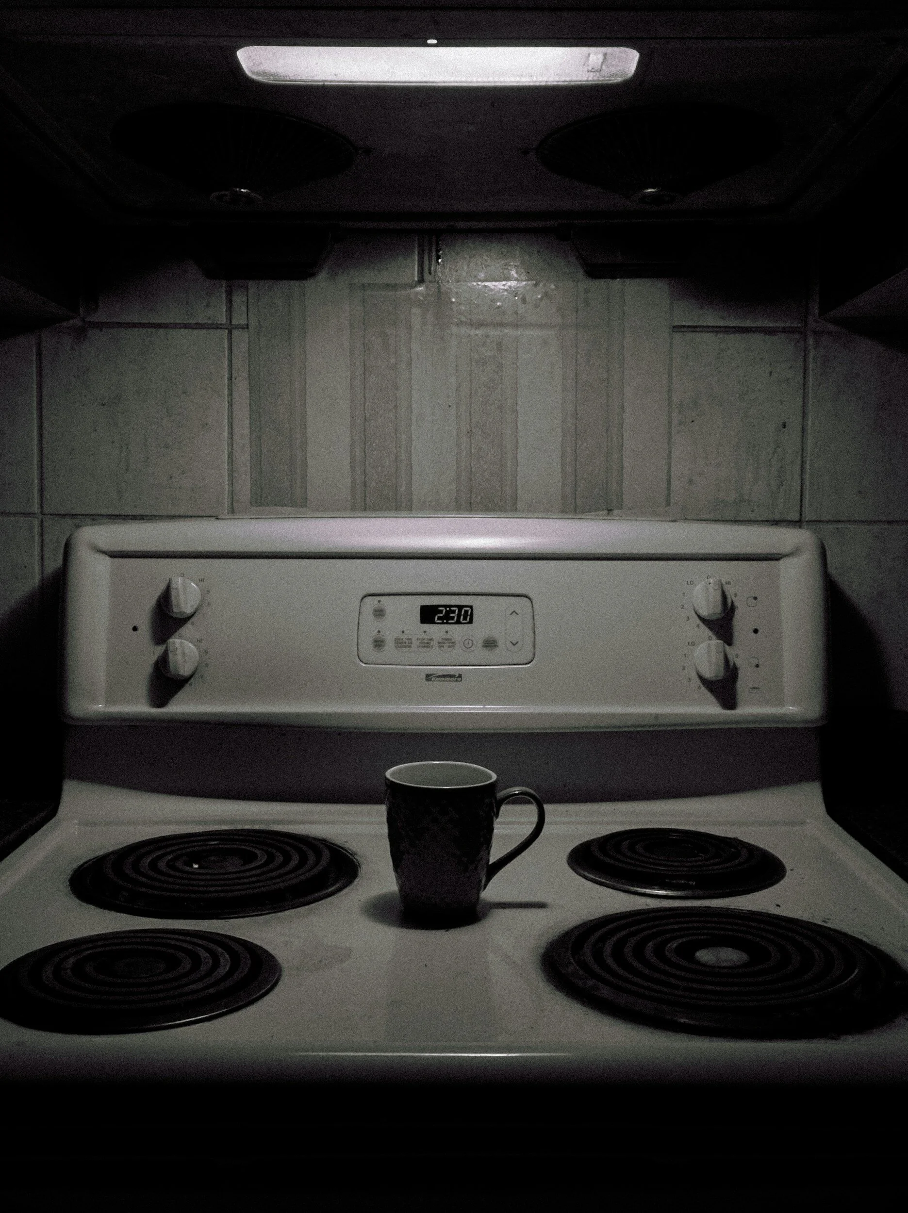A black coffee mug on a white electric stovetop with four burners, behind which is a white oven with control panel and timer set at 2:30, and a tiled wall in the background.