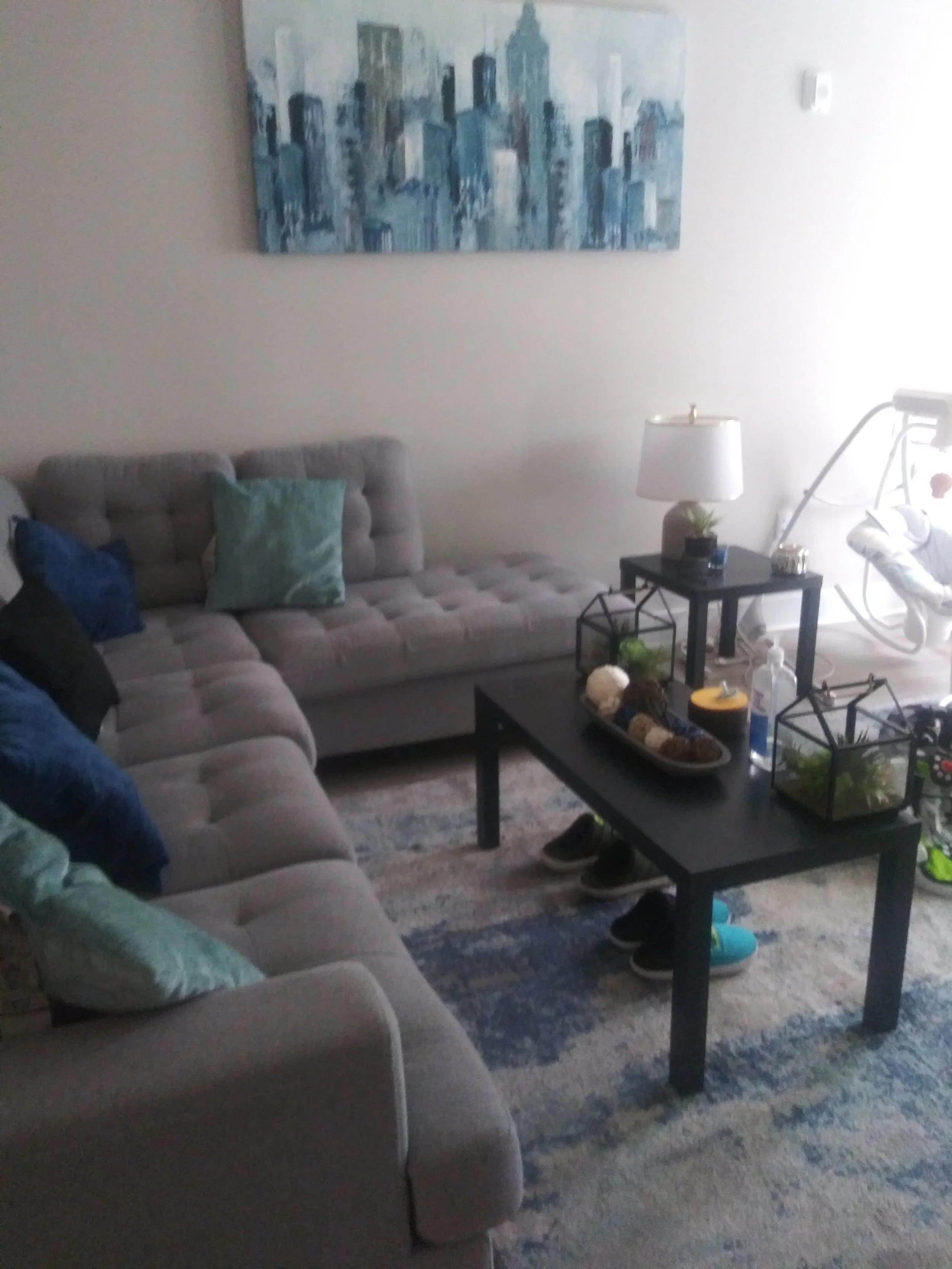Living room with gray sectional sofa, throw pillows, black coffee table with decor, side table with lamp, wall art of cityscape, and baby swing.