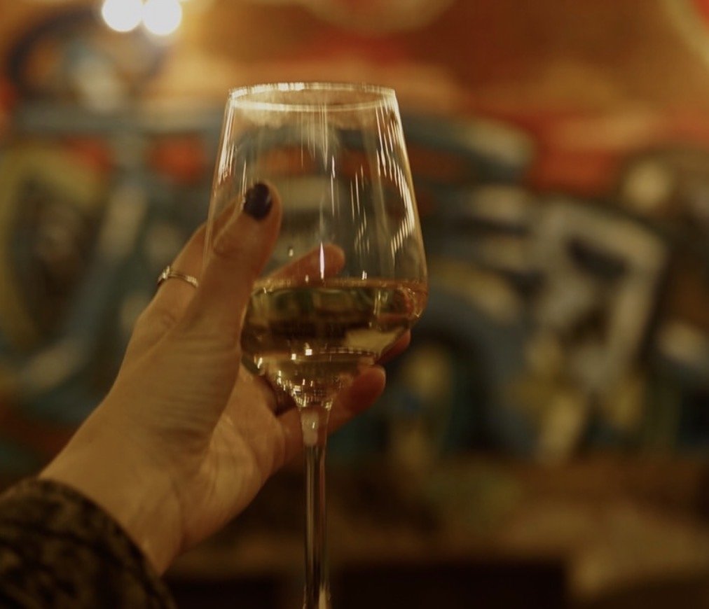 A hand holding a glass of white wine in a dimly lit setting with colorful, abstract graffiti art in the background.