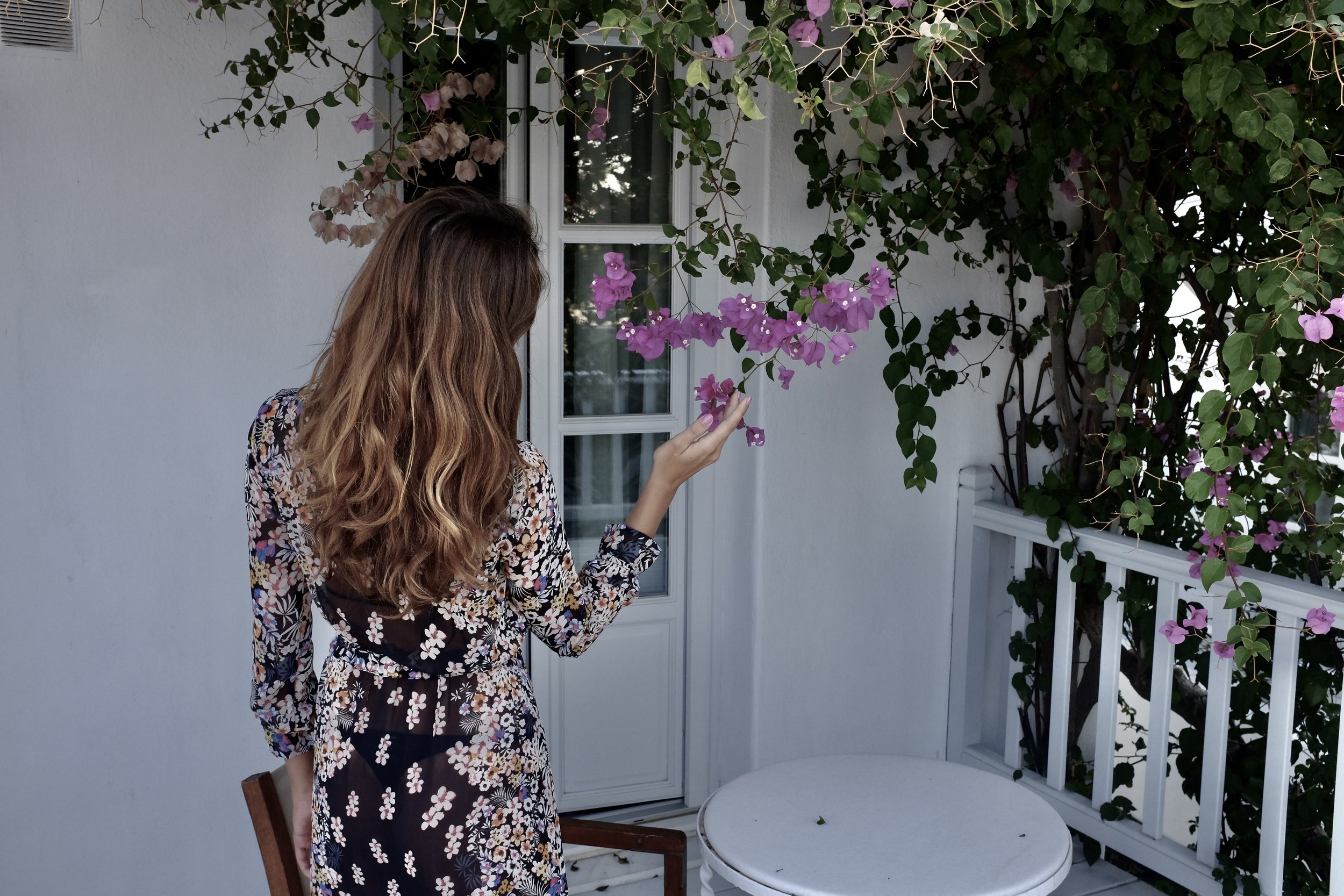 Dostinja in floral dress facing a white door, holding pink bougainvillea flowers, with greenery around.
