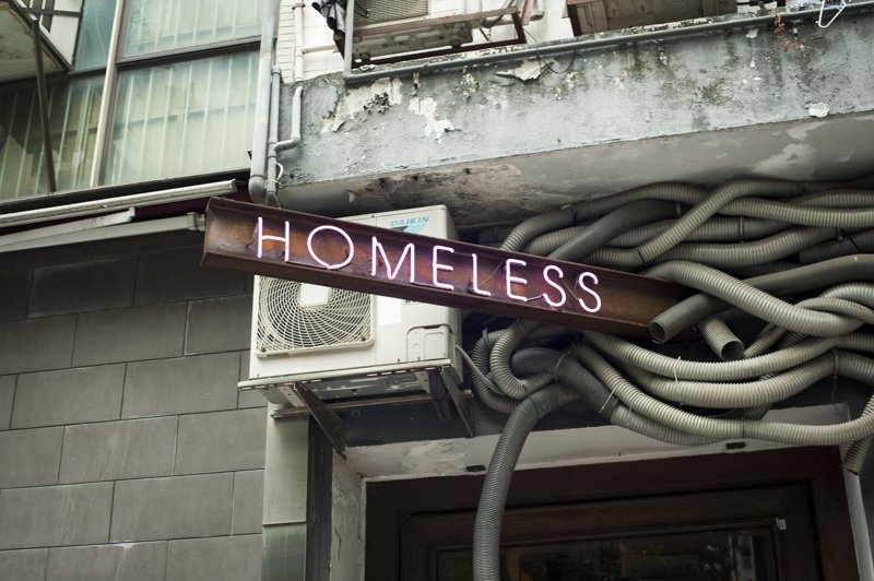 Sign reading "HOMELESS" in white neon lights mounted on a building exterior with visible air conditioner and exposed cables.