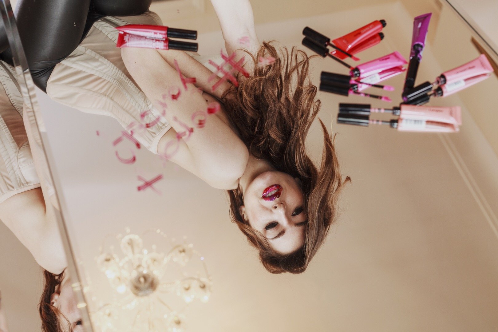 Dostinja looking in mirror with various lip glosses and writing on glass surface.