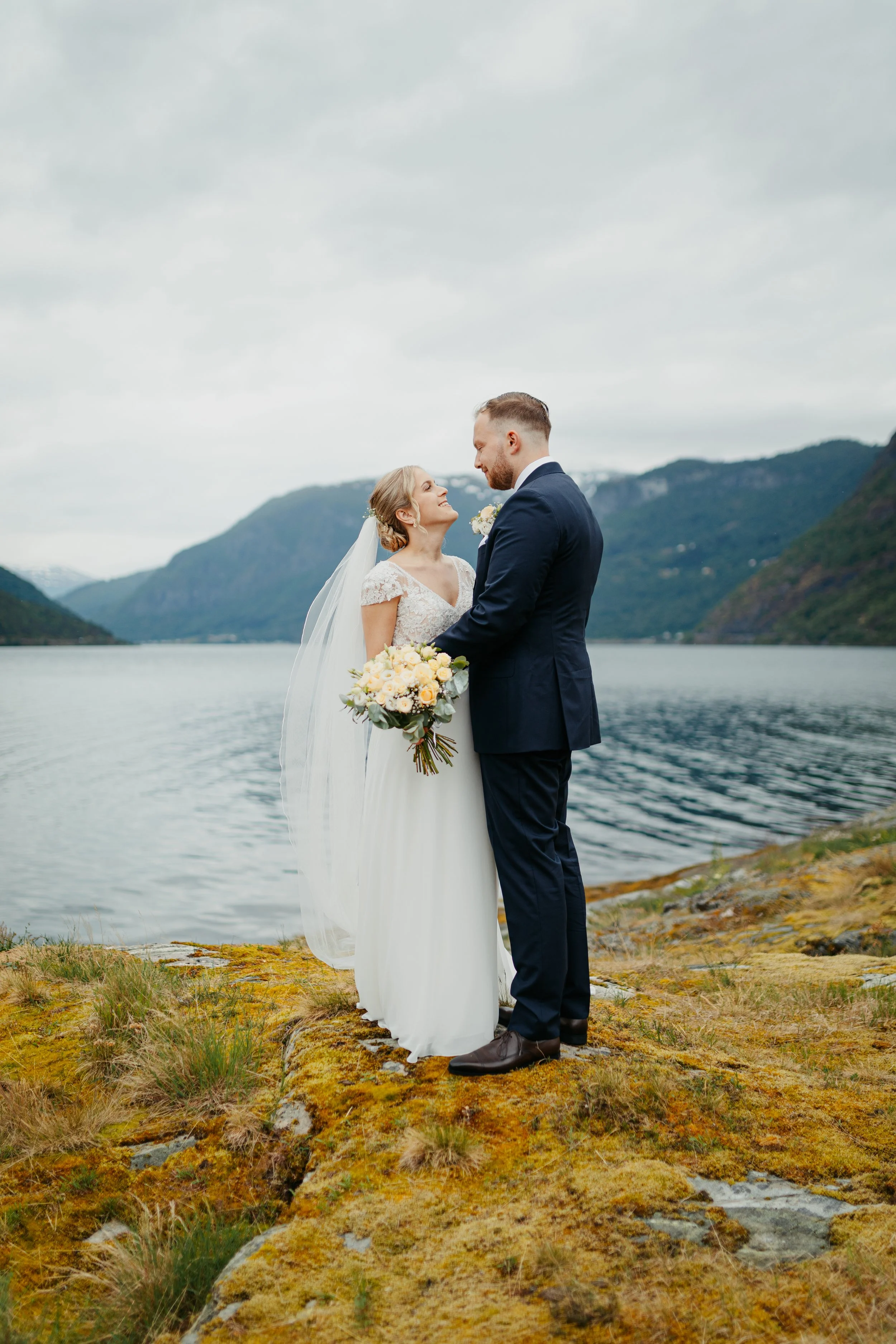Et kyssende brudepar står på en mosekledd klippe ved en innsjø, omgitt av fjell under en overskyet himmel.