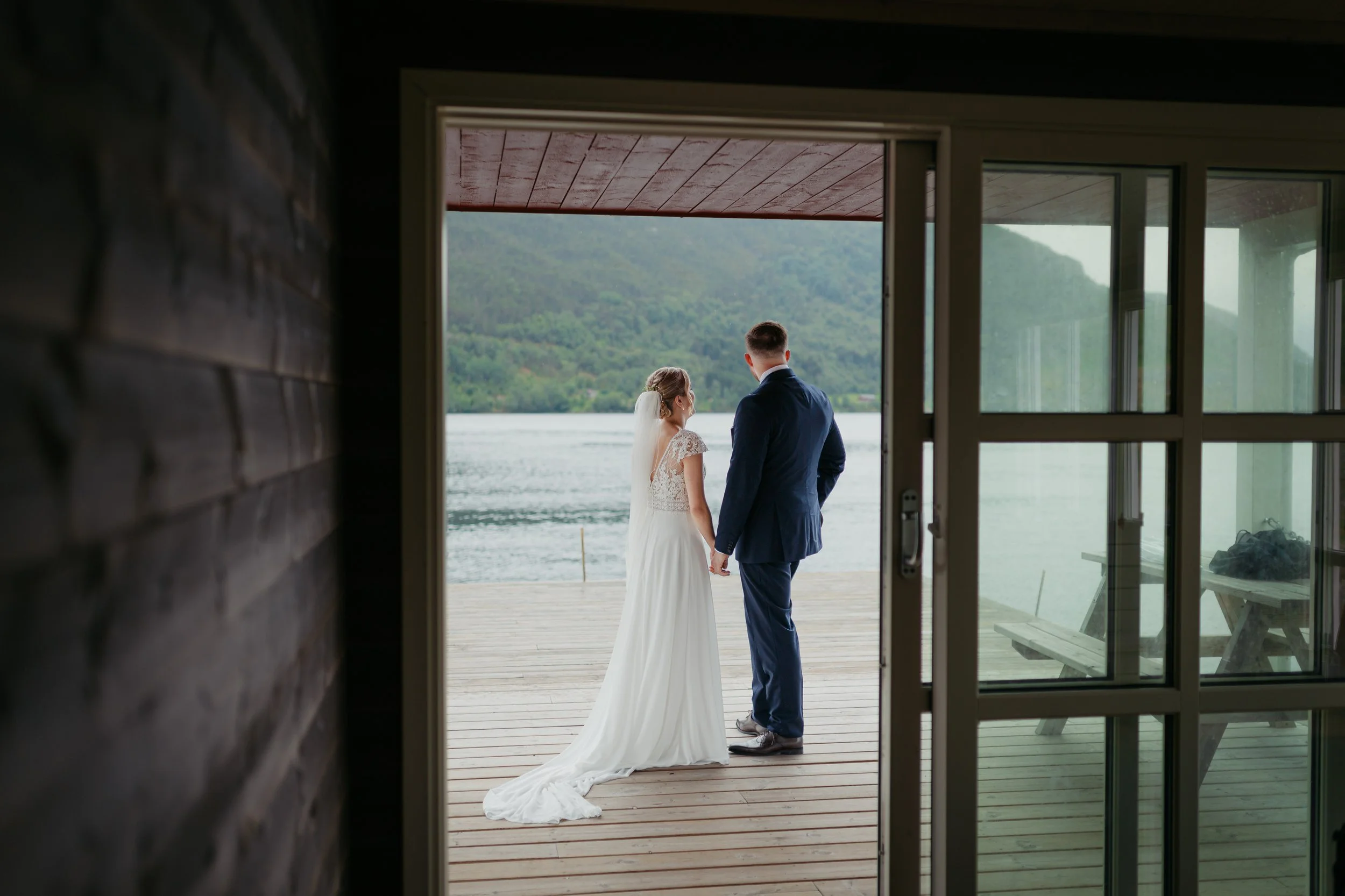 Et brudepar står på en brygge ved en innsjø, sett innendørs gjennom en glassdør.