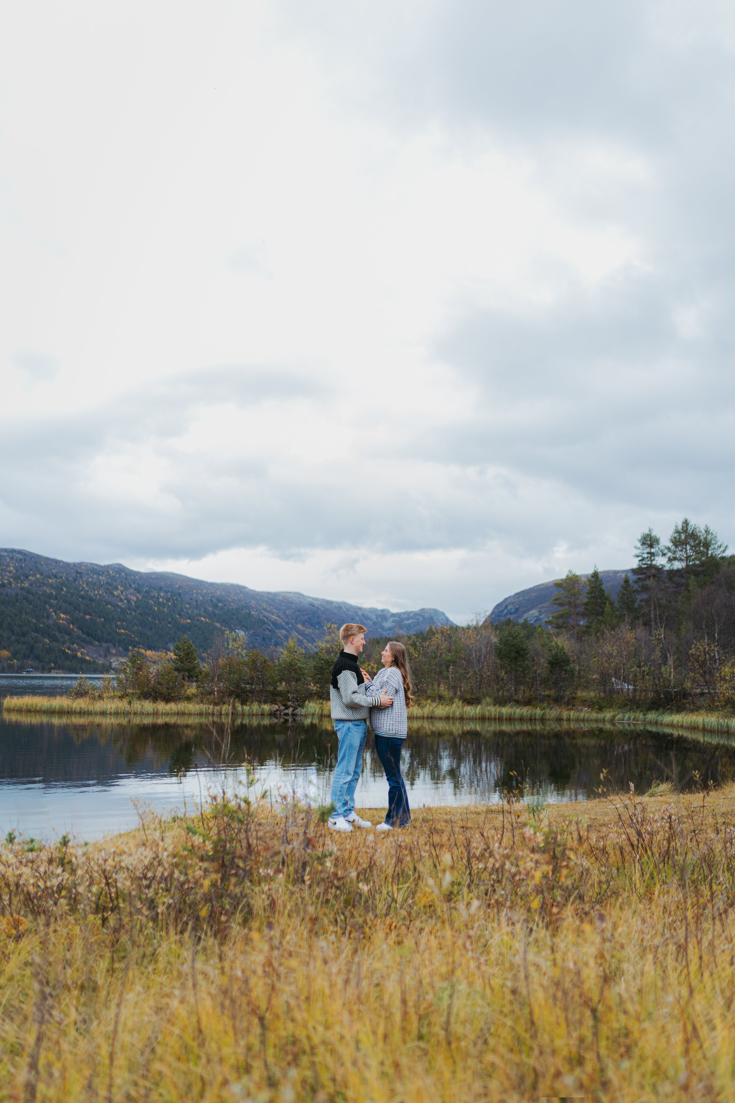 Et par står foran en innsjø omgitt av fjell og trær, og ser på hverandre.