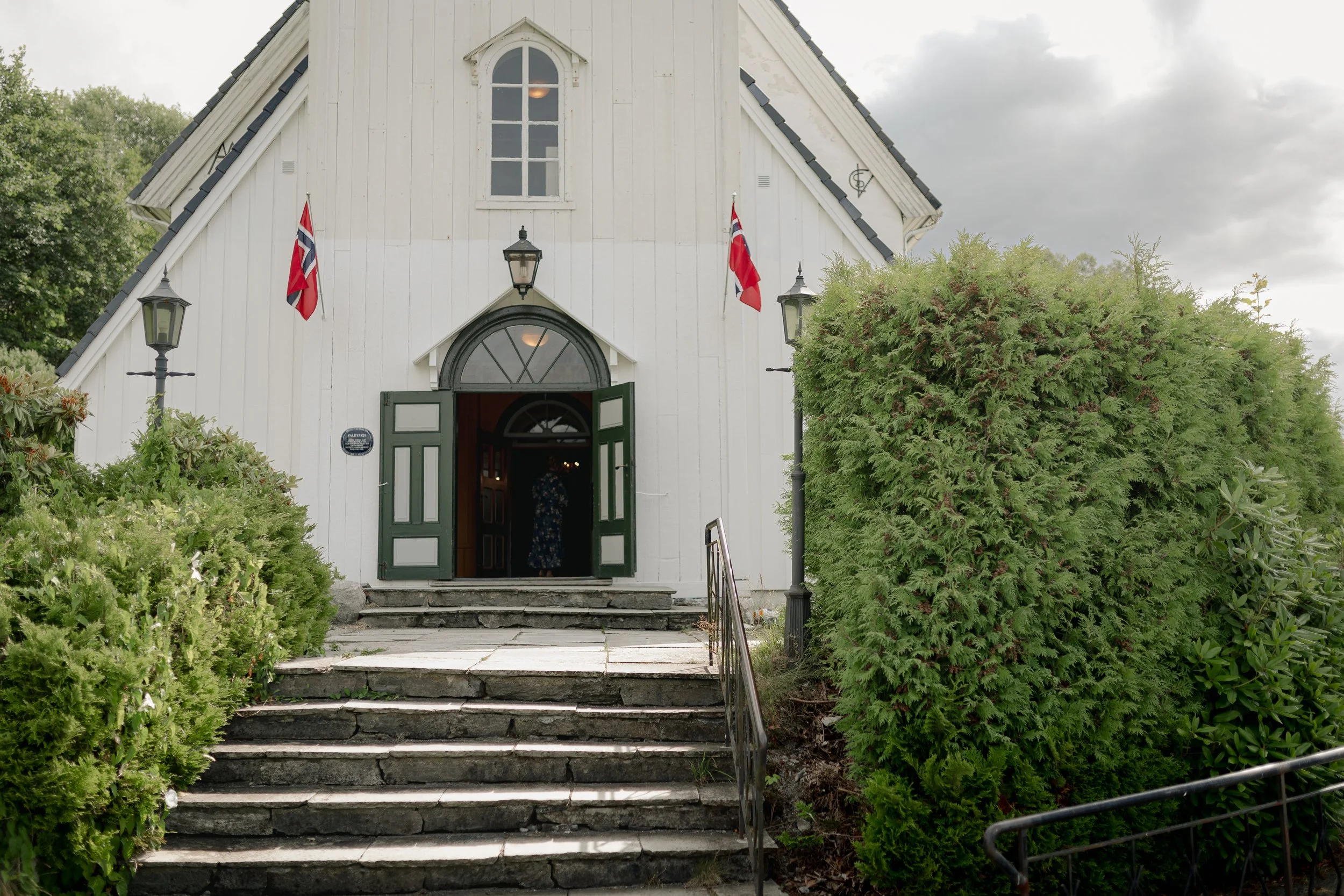 En hvit kirke med åpen dør, omgitt av grønne busker og lamper, med norske flagg på hver side av inngangen.