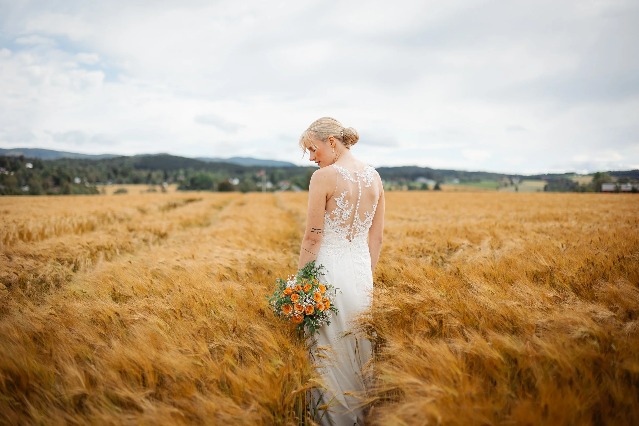 En brud i hvit brudekjole står i en gylden kornåker og holder en blomsterbukett.