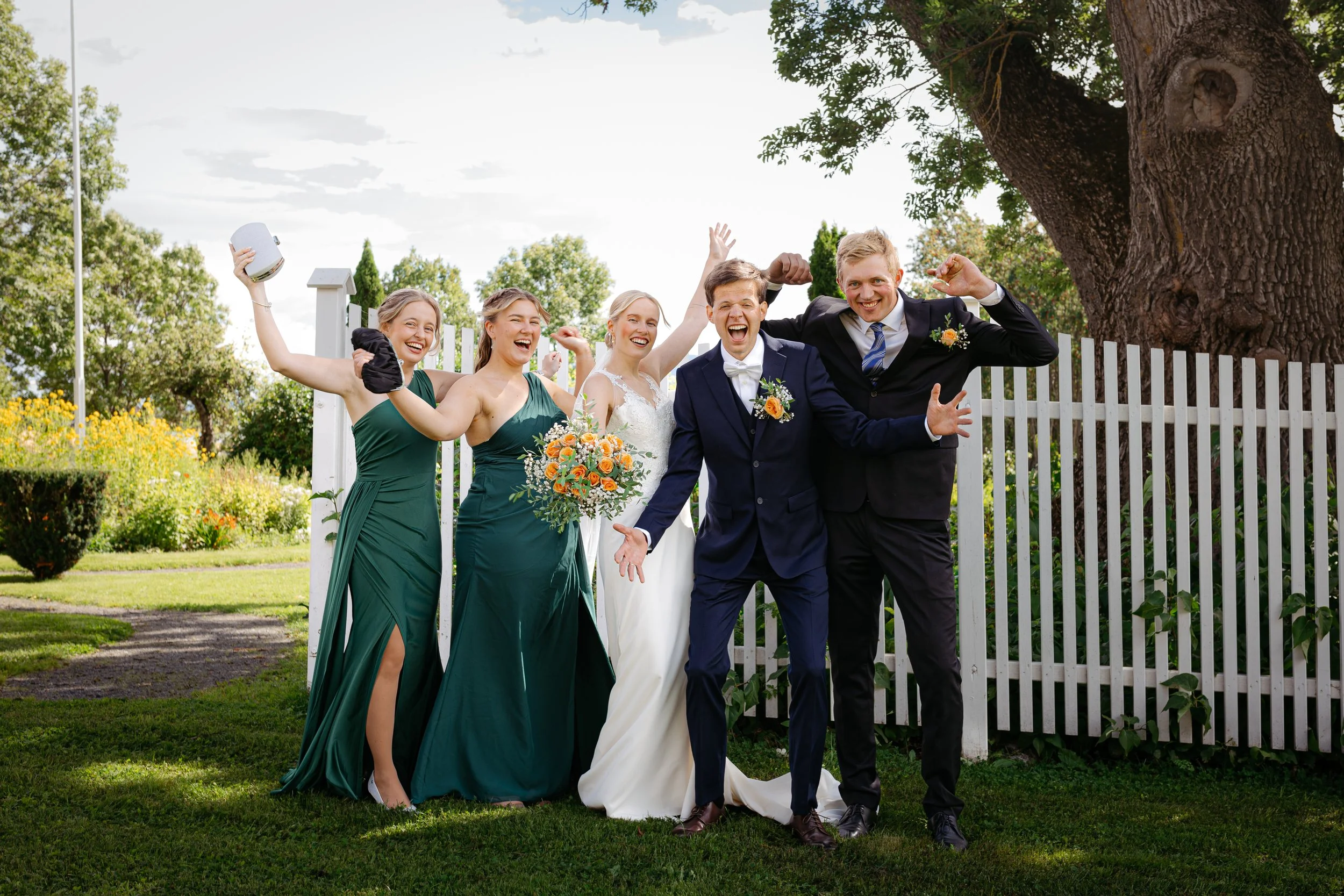 Gruppe av mennesker i bryllupsantrekk ute i hagen, feirer og smiler.