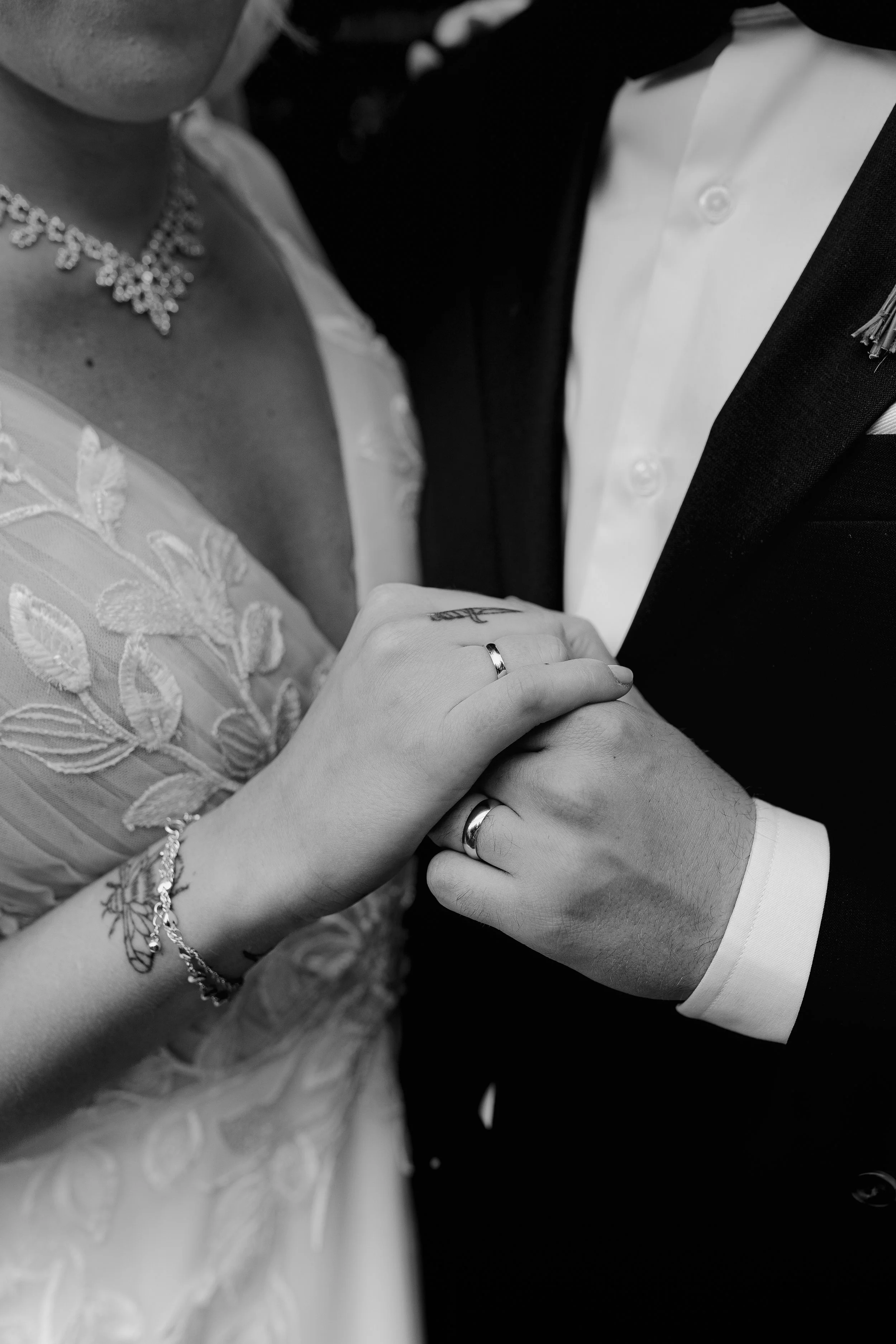 Nærbilde av et brudepar som holder hender, i svart-hvitt, fokus på bryllingsringene, med del av brudens hånd, kjole og smykke, og del av den grooms smoking.