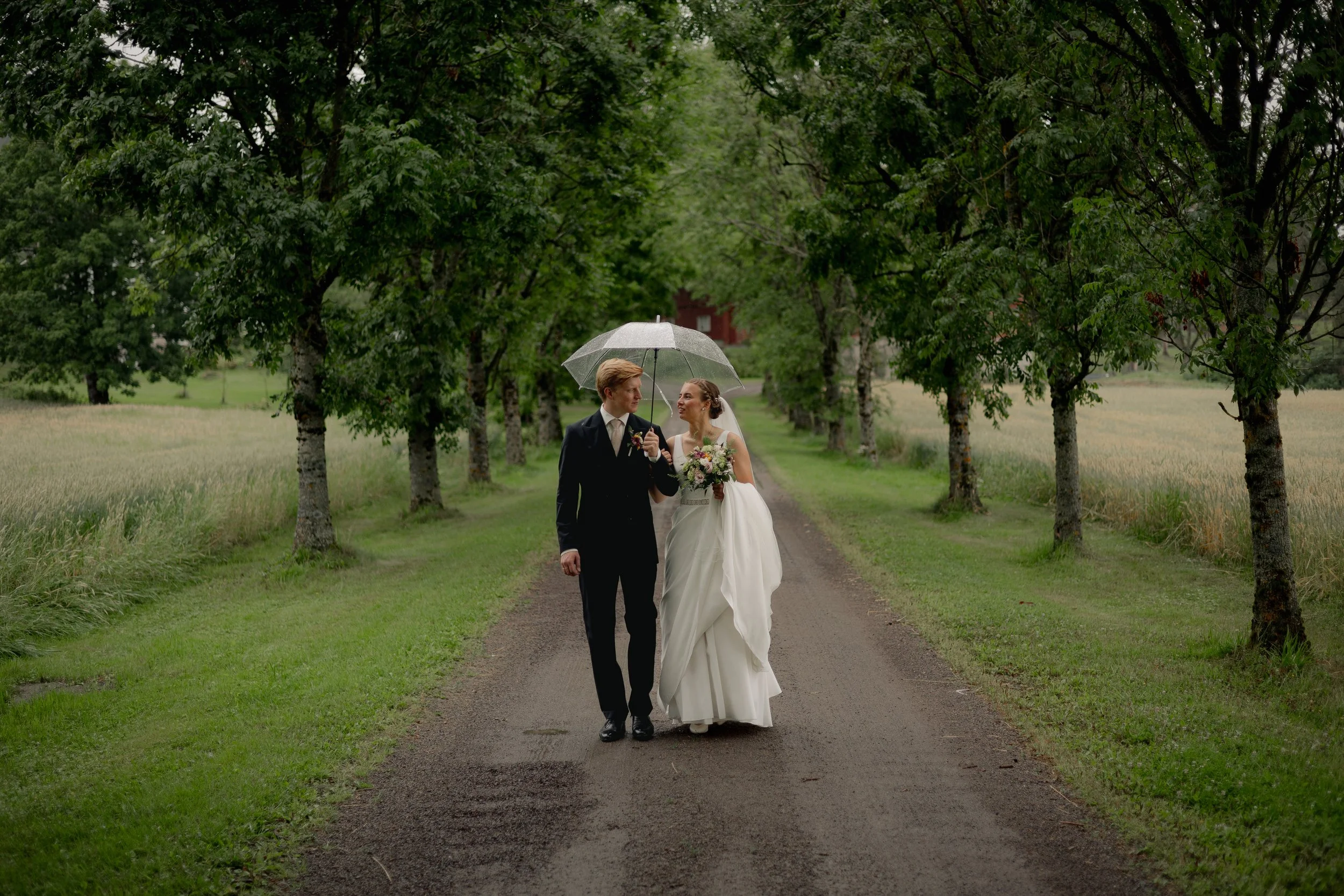Et nygifte par går hånd i hånd under et transparent paraply i en grønn, trivelig allé med trær på begge sider.