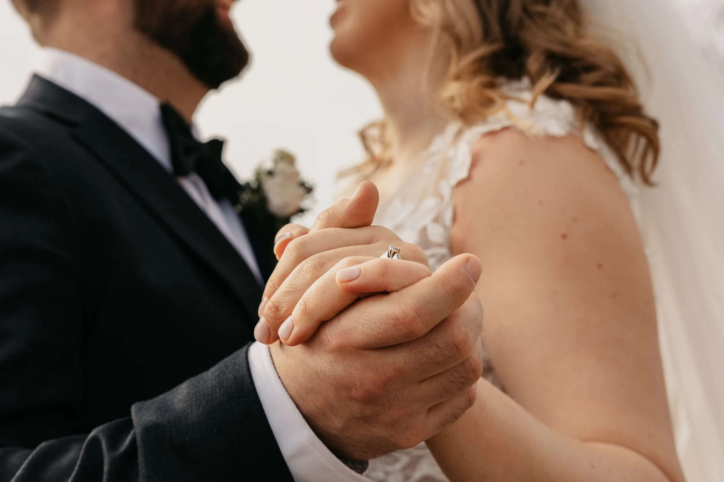 Nærbilde av et ektepar som holder hender, med fokus på forlovelsesringen, i bryllupsklær.