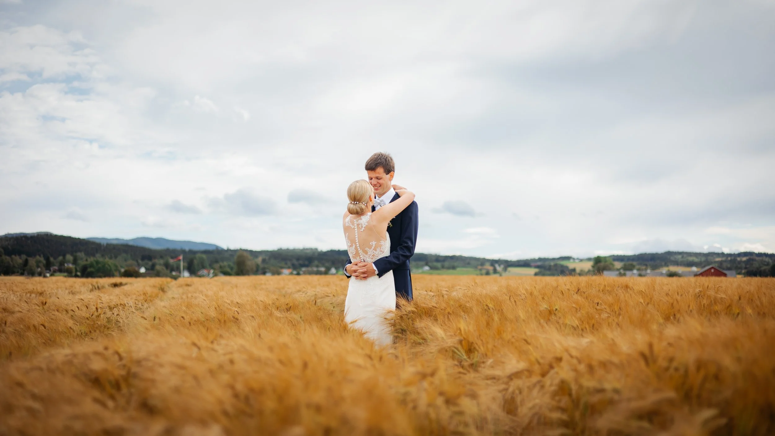 En brud og en brudgom som omfavner i en åker med gyllen korn. Himmel med skyer i bakgrunnen.