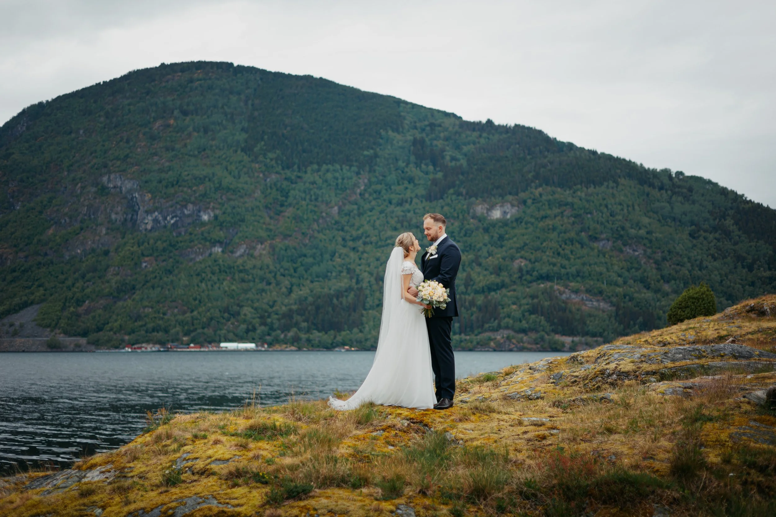 Et brudepar står på en mosekledd landtunge ved en innsjø med en stor, grønn fjell i bakgrunnen.