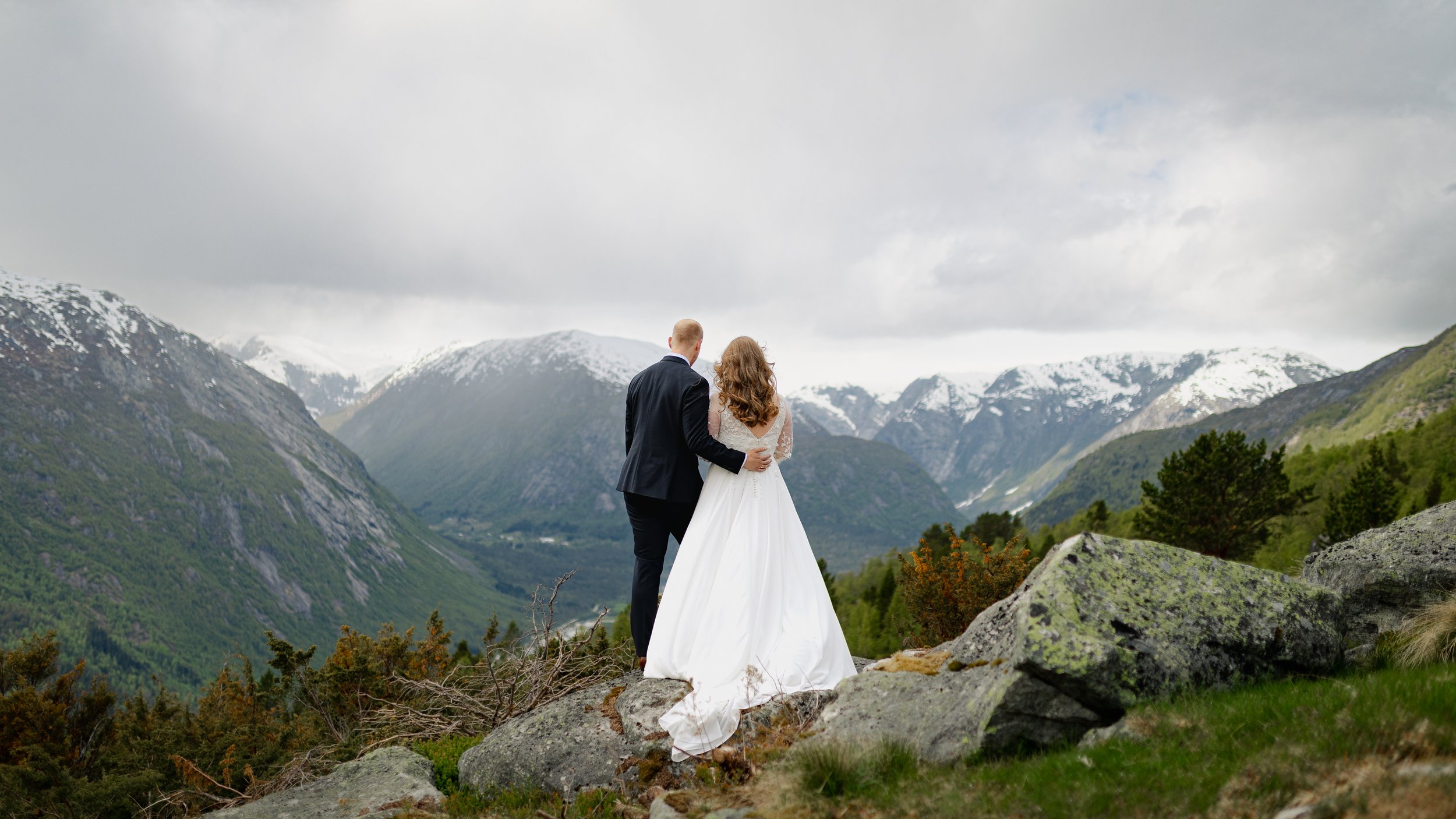Et brudepar står på en fjellutsikt med snødekte fjell i bakgrunnen, dress og brudekjole.
