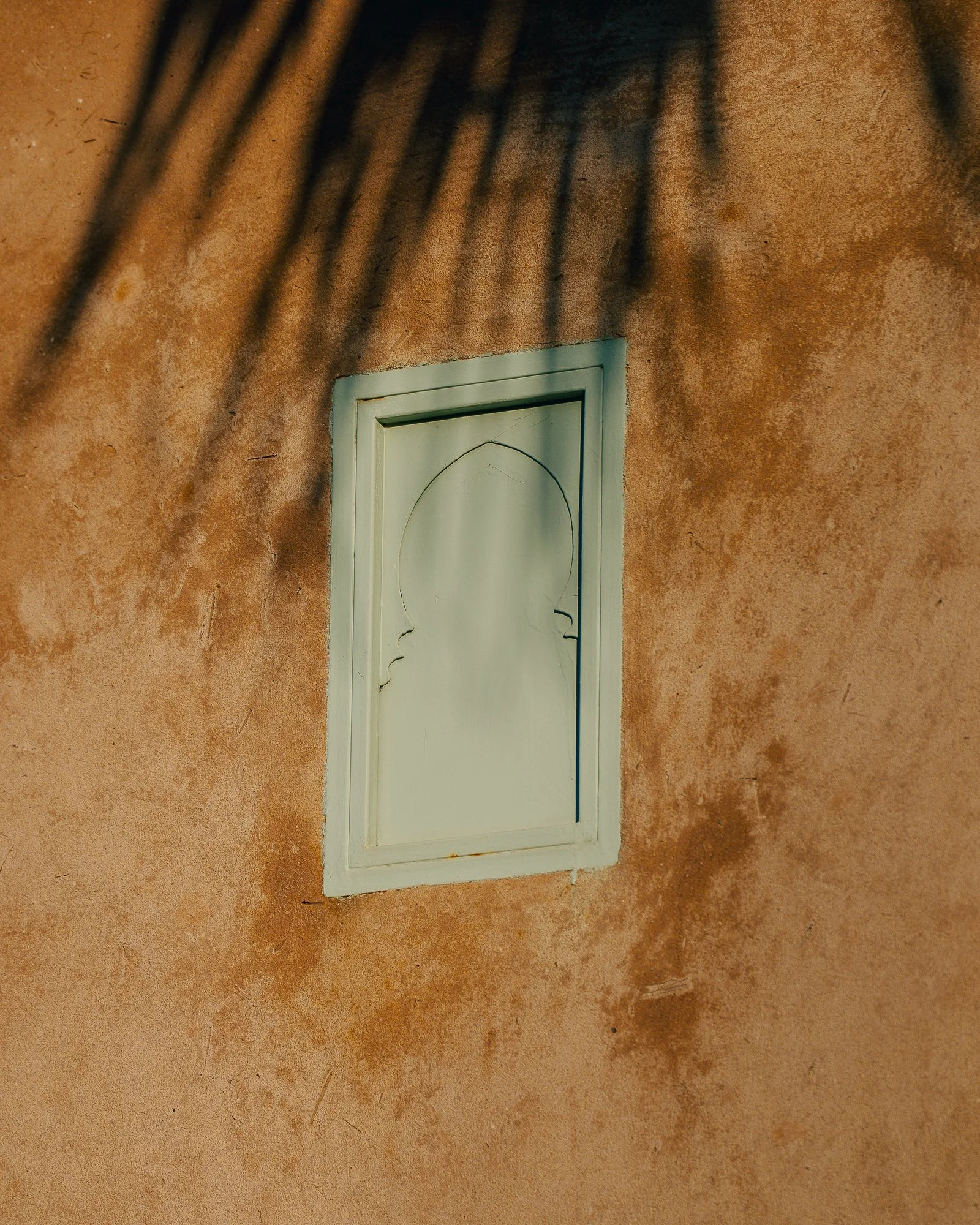 A white rectangular window with decorative sides on a textured reddish-brown wall, with dark shadows from plant leaves at the top.