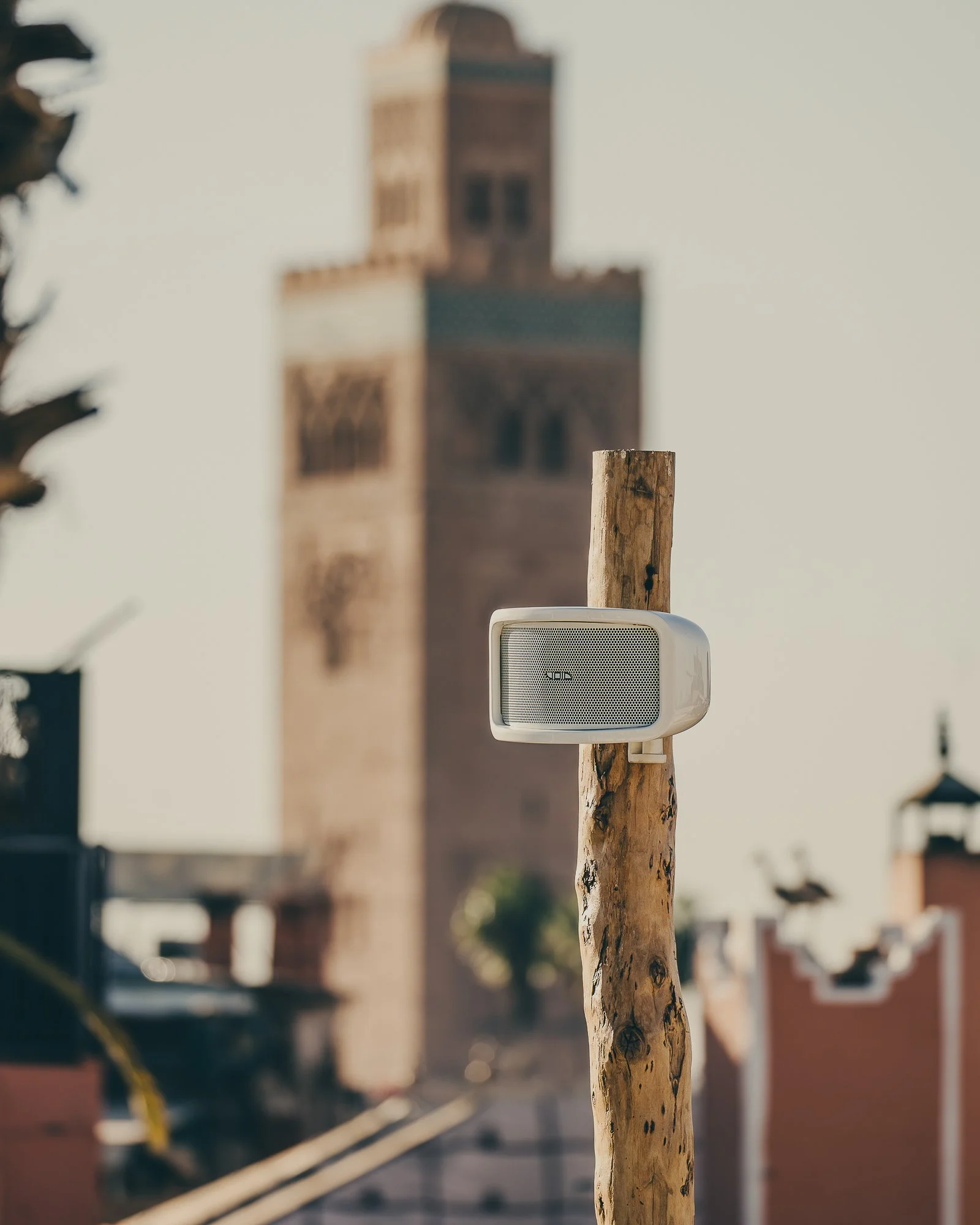 A white speaker mounted on a wooden post with the blurred background of a cityscape including a tall tower.