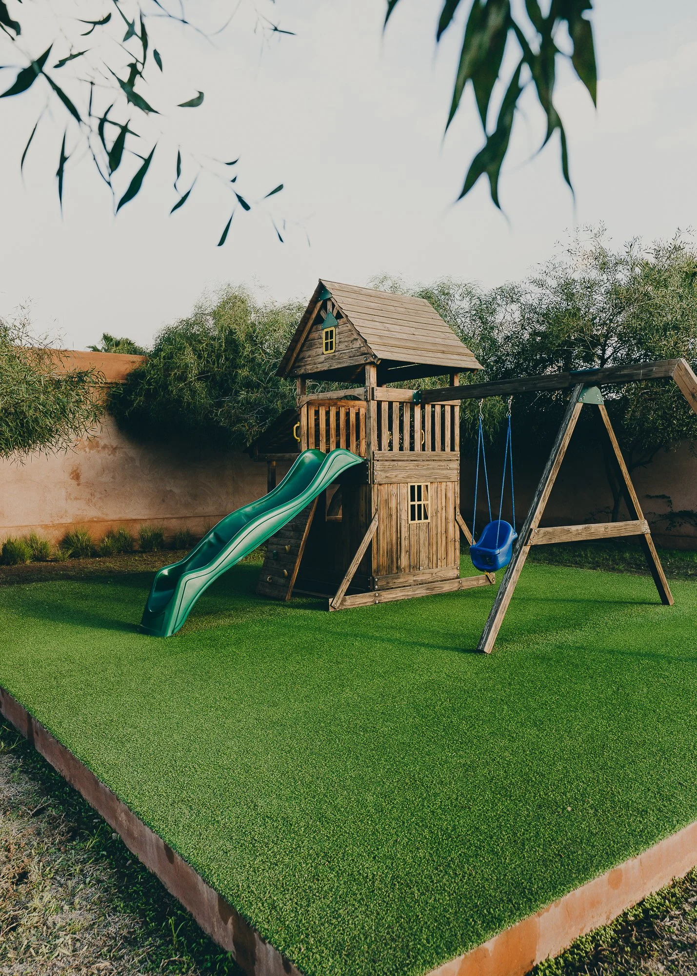 A backyard playground with a wooden house structure, green slide, and blue swing set on a grassy surface, with trees and a concrete wall in the background.