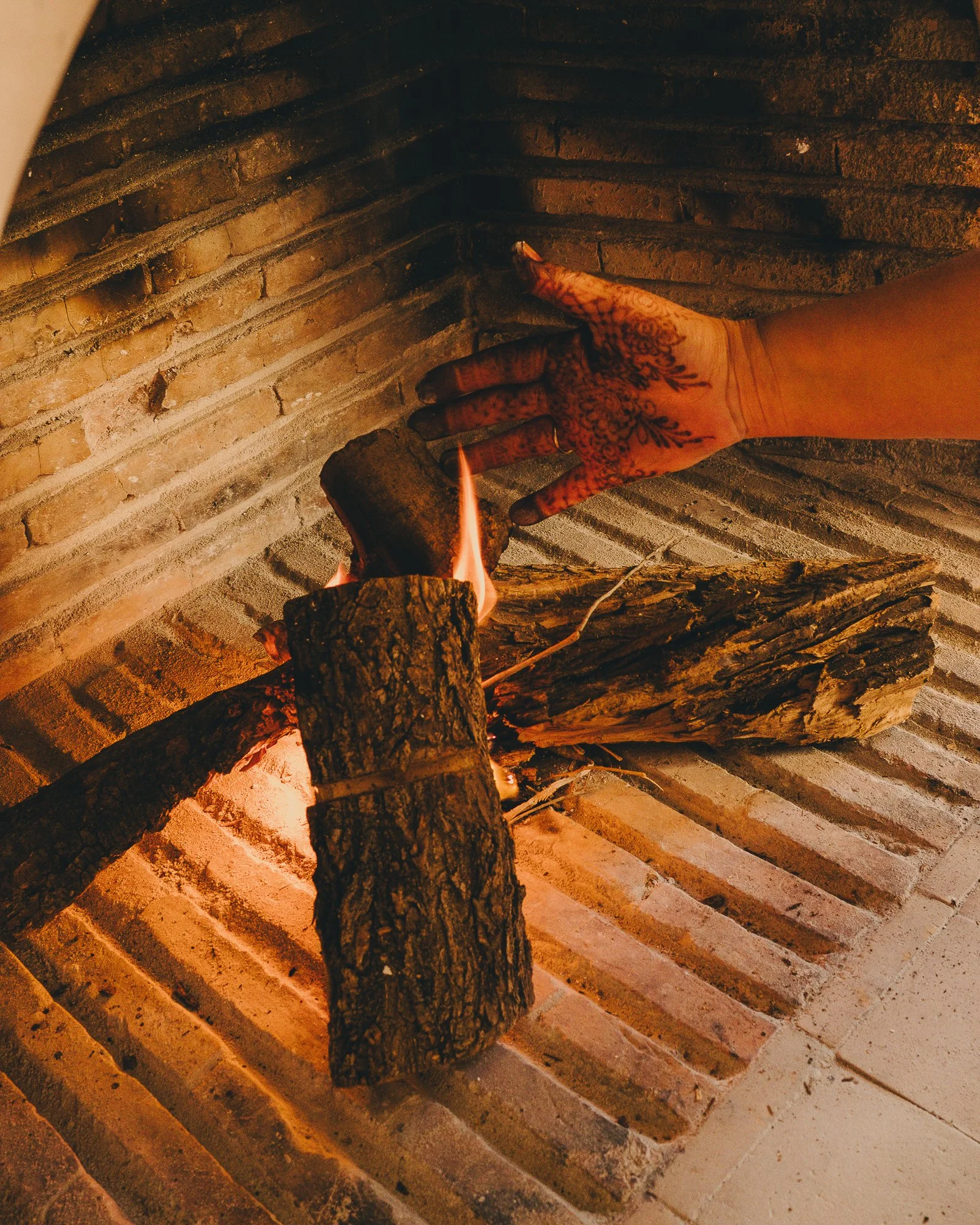Person with henna tattoos on their hand placing a small piece of wood onto a fire inside a brick fireplace.