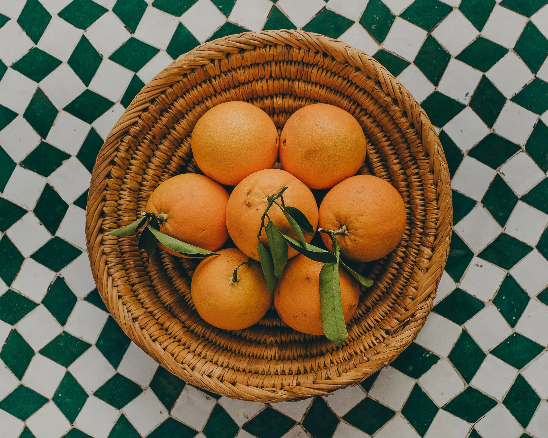 A wicker basket filled with seven oranges, some with green leaves, placed on a green and white diamond-patterned tile surface.
