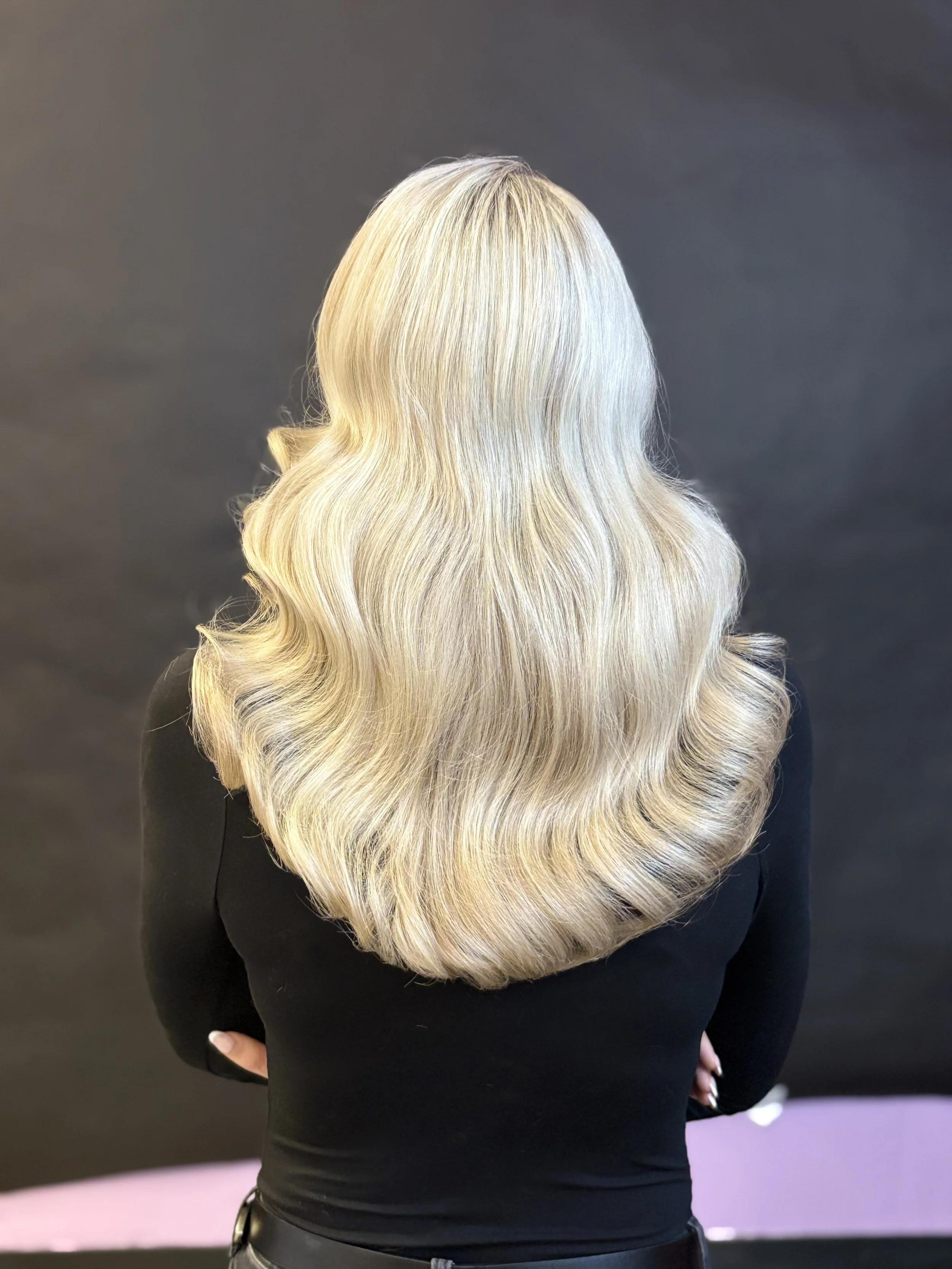 Back view of a woman with long, wavy, blonde hair against a dark background. Blonde Highlights.