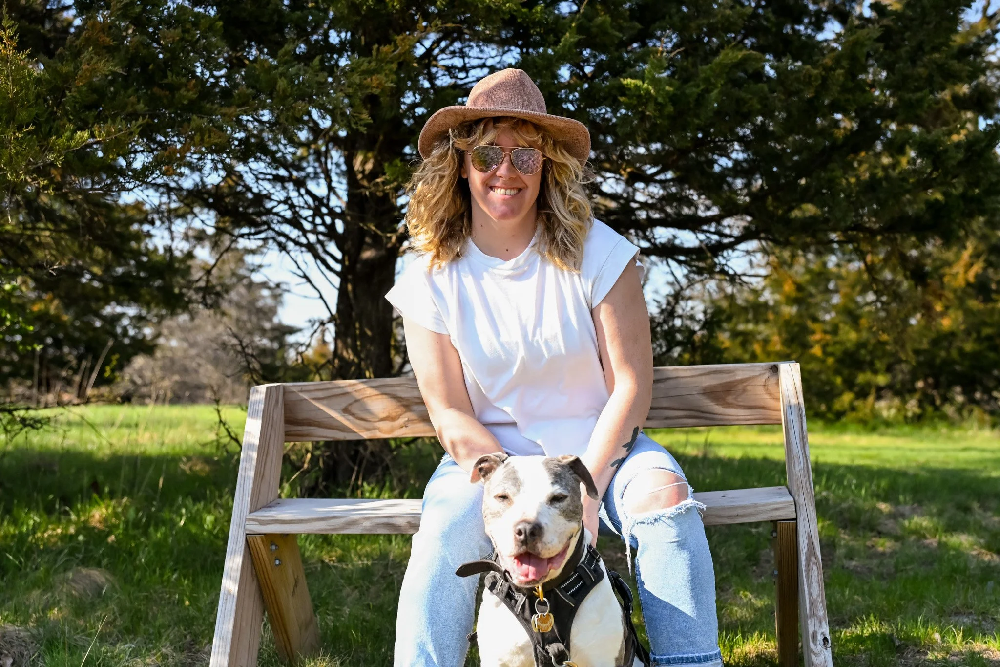 A woman with curly blonde hair wearing sunglasses, a beige wide-brim hat, and a white t-shirt, sitting on a wooden bench outdoors with a smiling dog in her lap, in front of a background of trees and green grass.